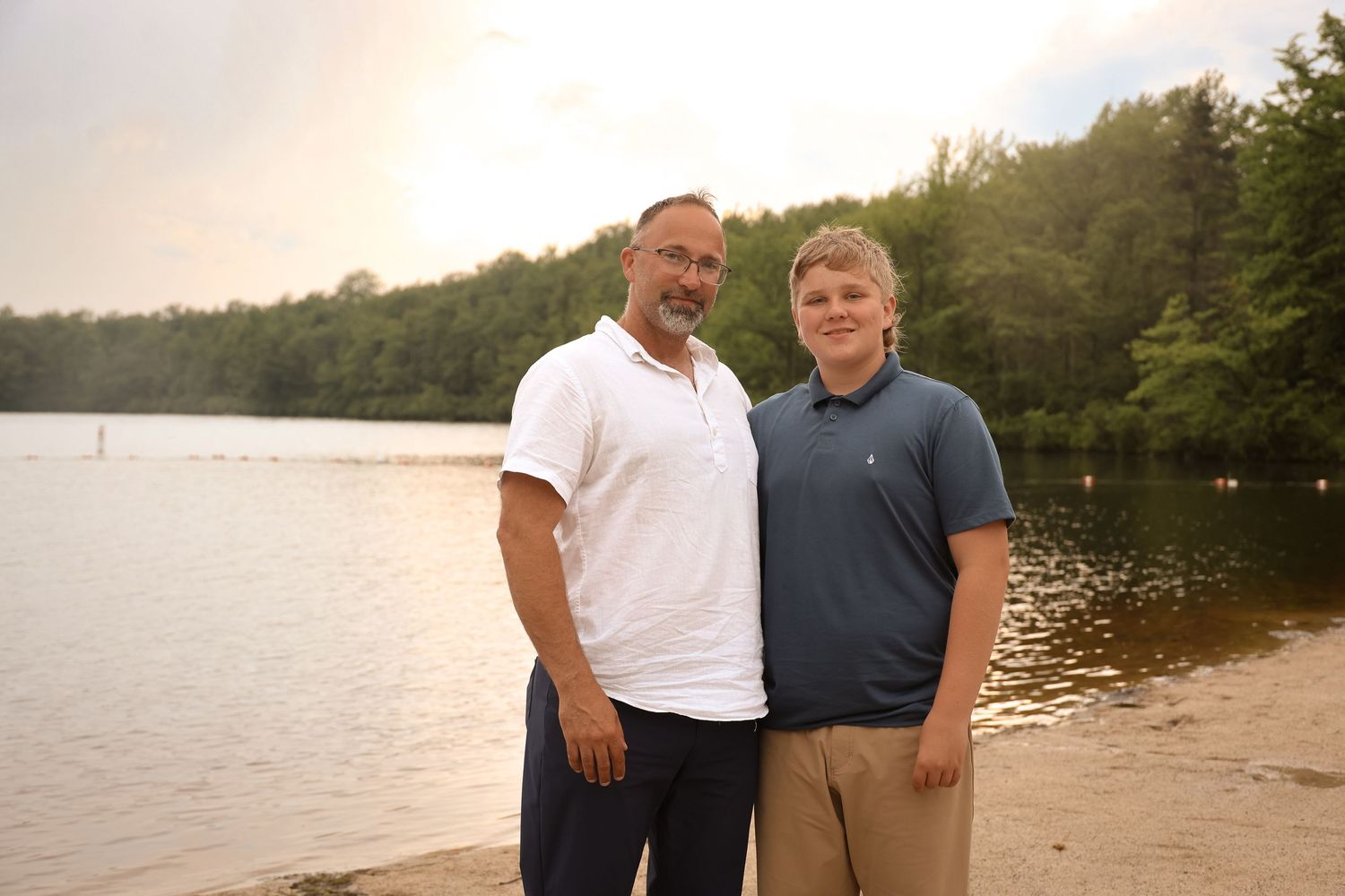 Two people stand close together on a beach with a tranquil lake and wooded shoreline behind them.