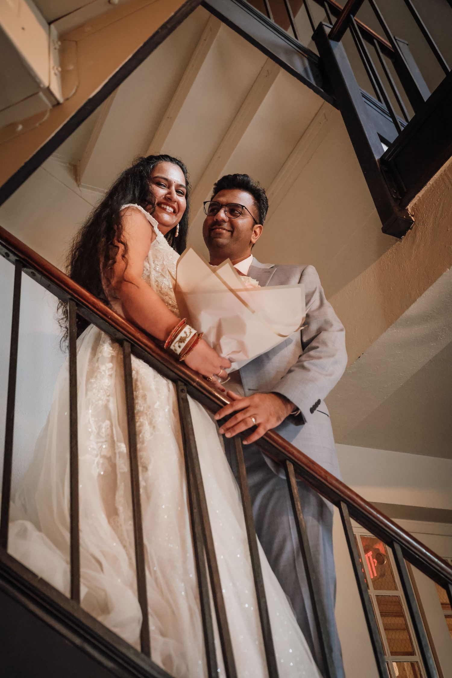 A couple stands together on a staircase, the bride wearing a white lace dress and groom in light colored formal attire.