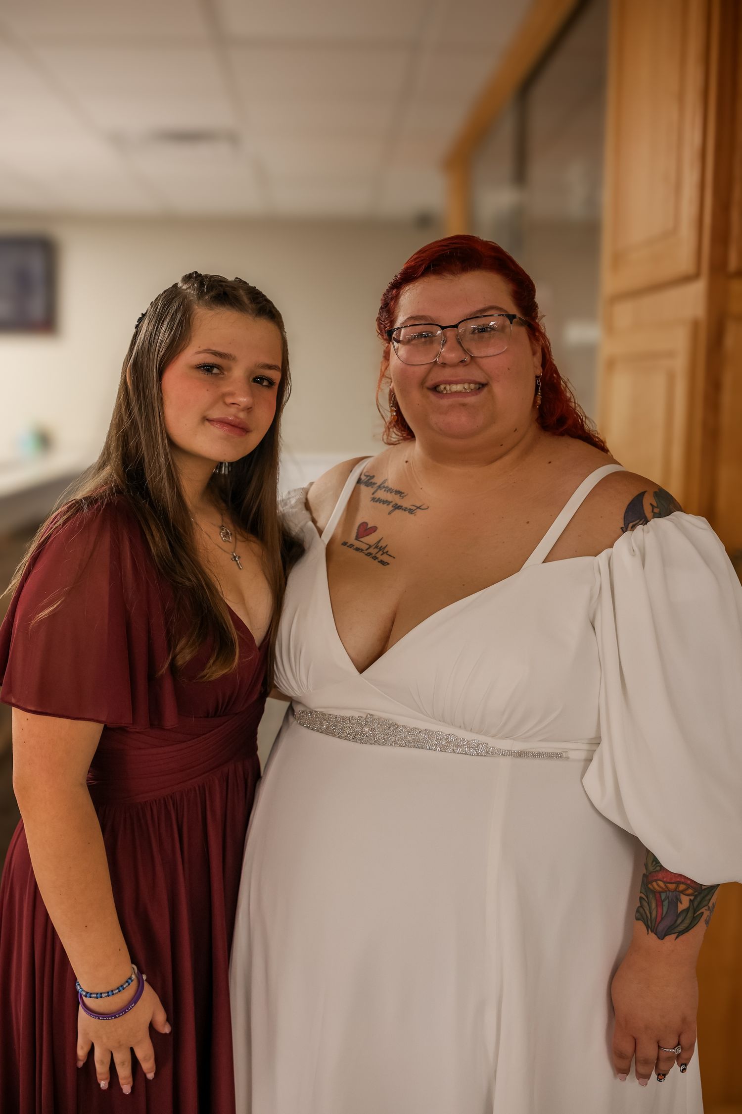 Two people pose together at a formal event, one wearing a burgundy dress and one in a white wedding gown with sparkly details.