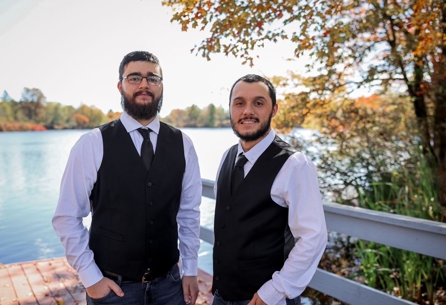A group of groomsmen in black vests and white shirts pose together outdoors during an autumn wedding celebration.