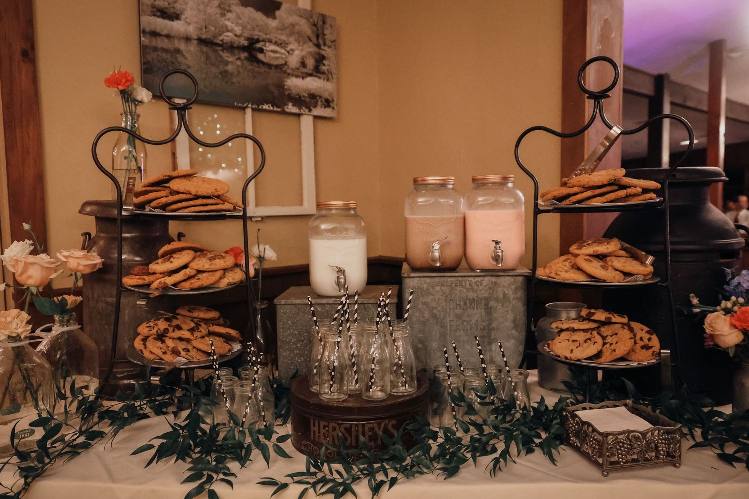 A rustic dessert display table with cookies on tiered stands, glass jars of milk, and greenery decorations.