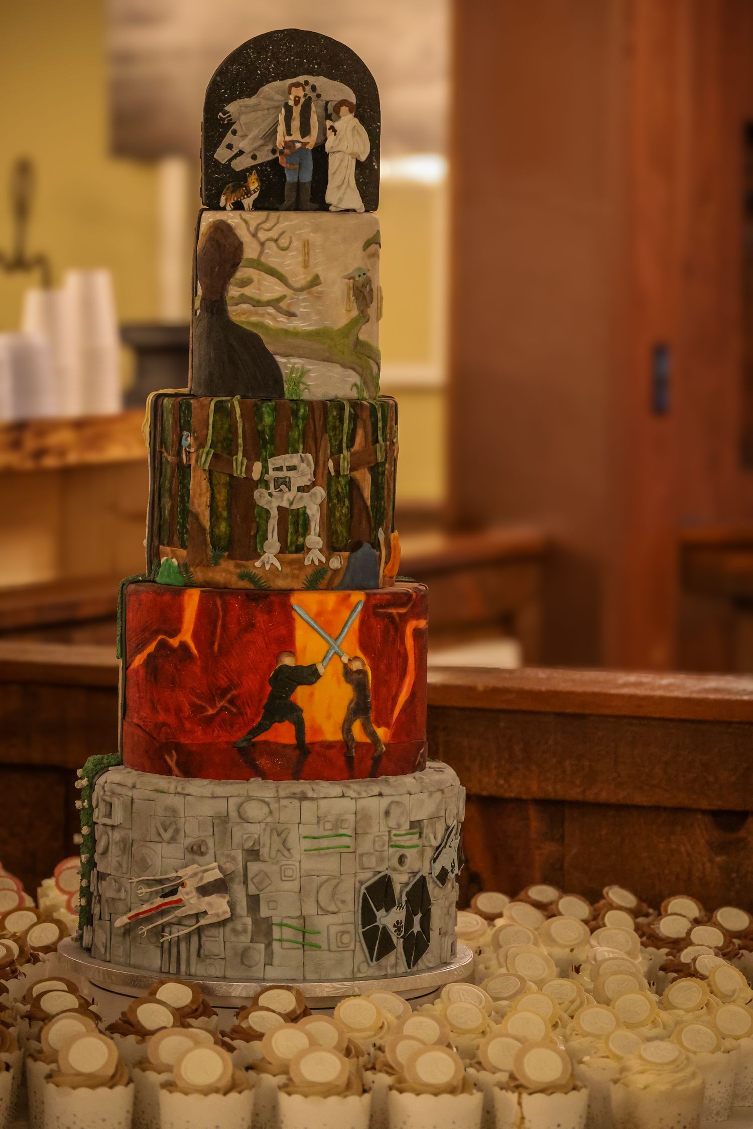 A multi-tiered artistic wedding cake decorated with silhouettes and scenes in different colors, surrounded by white cupcakes.