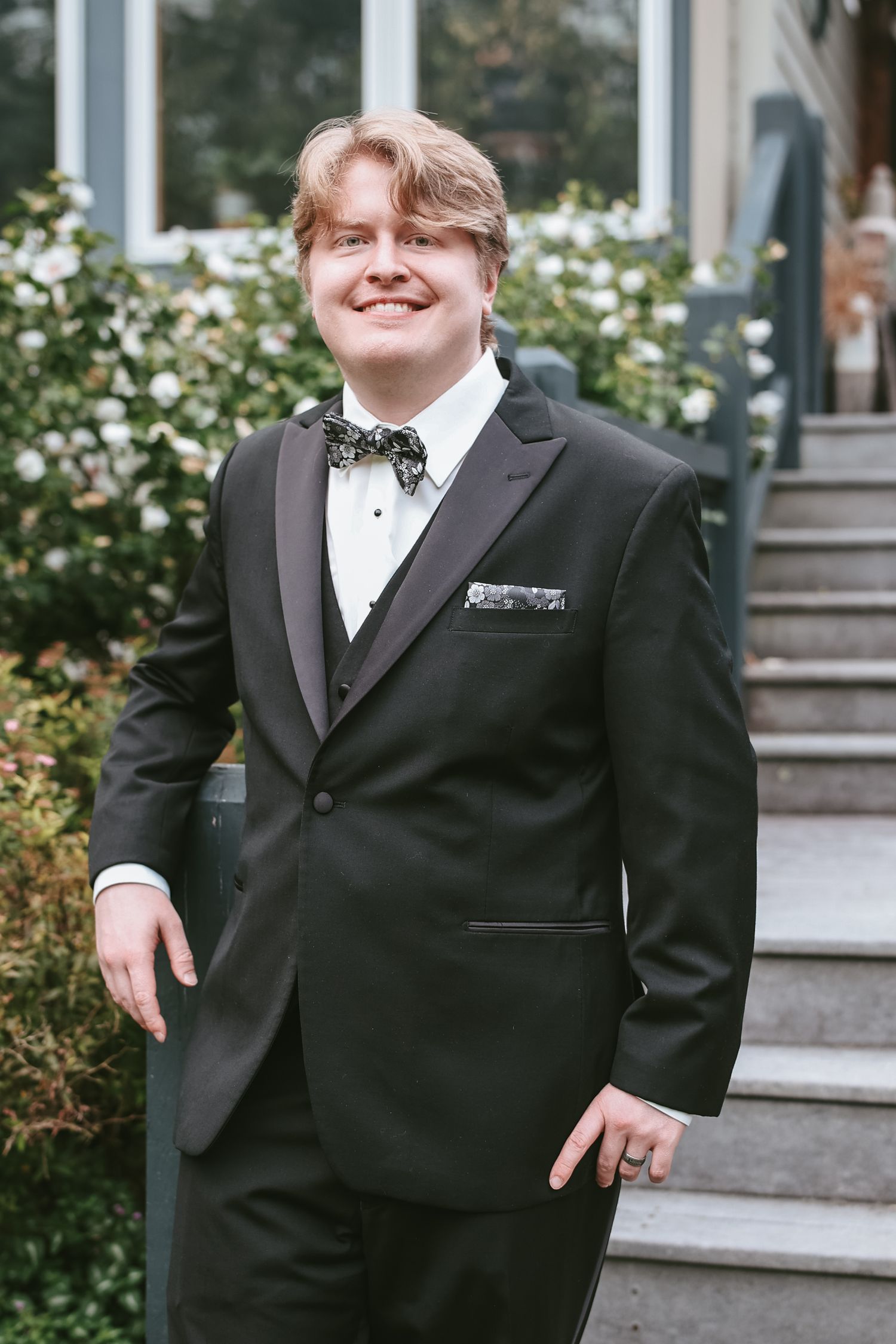 A person in a black tuxedo and bow tie poses on outdoor stone steps near white flowers and greenery.