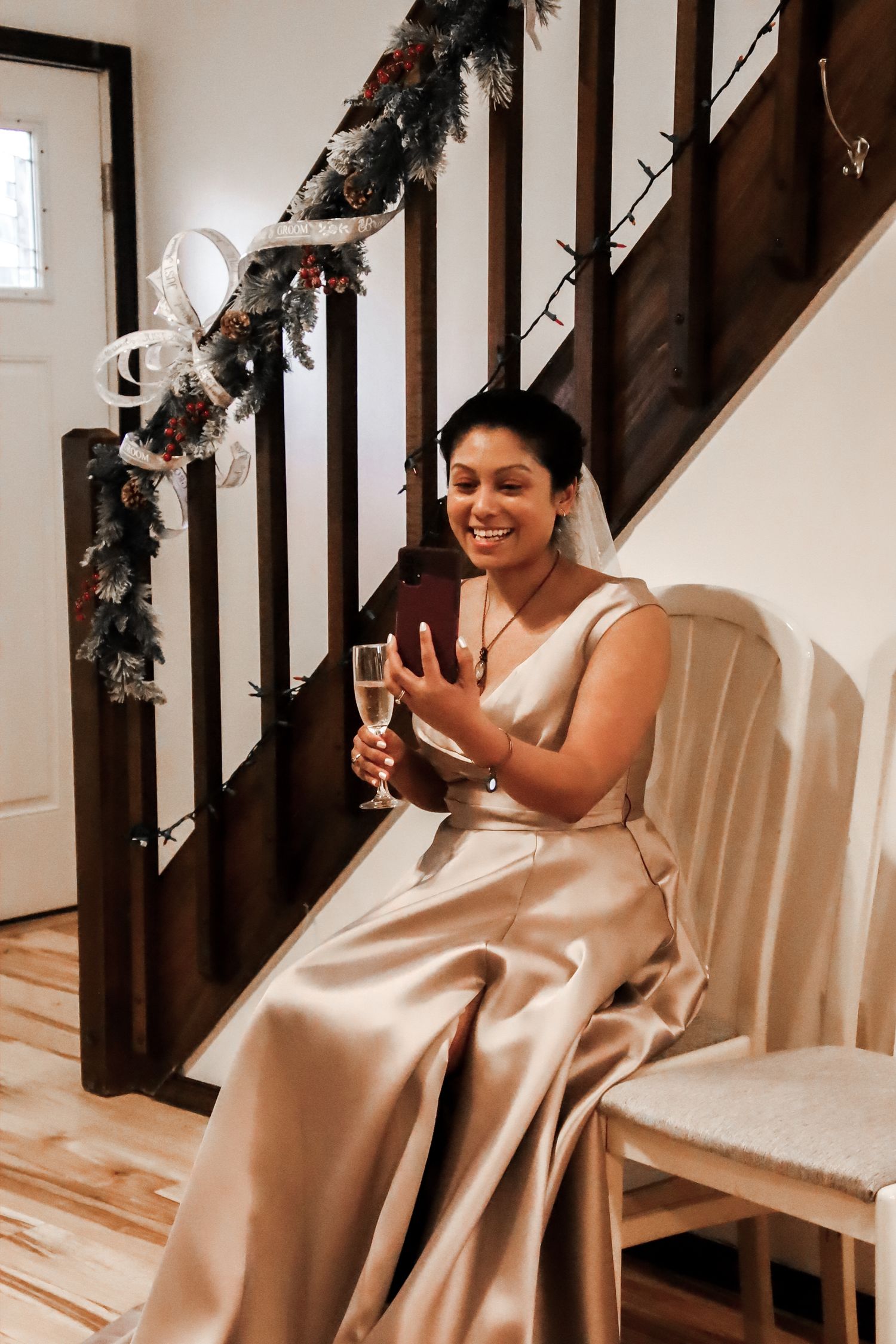 A person in a champagne-colored satin wedding dress sits on a chair by decorated stairs holding a glass of champagne.