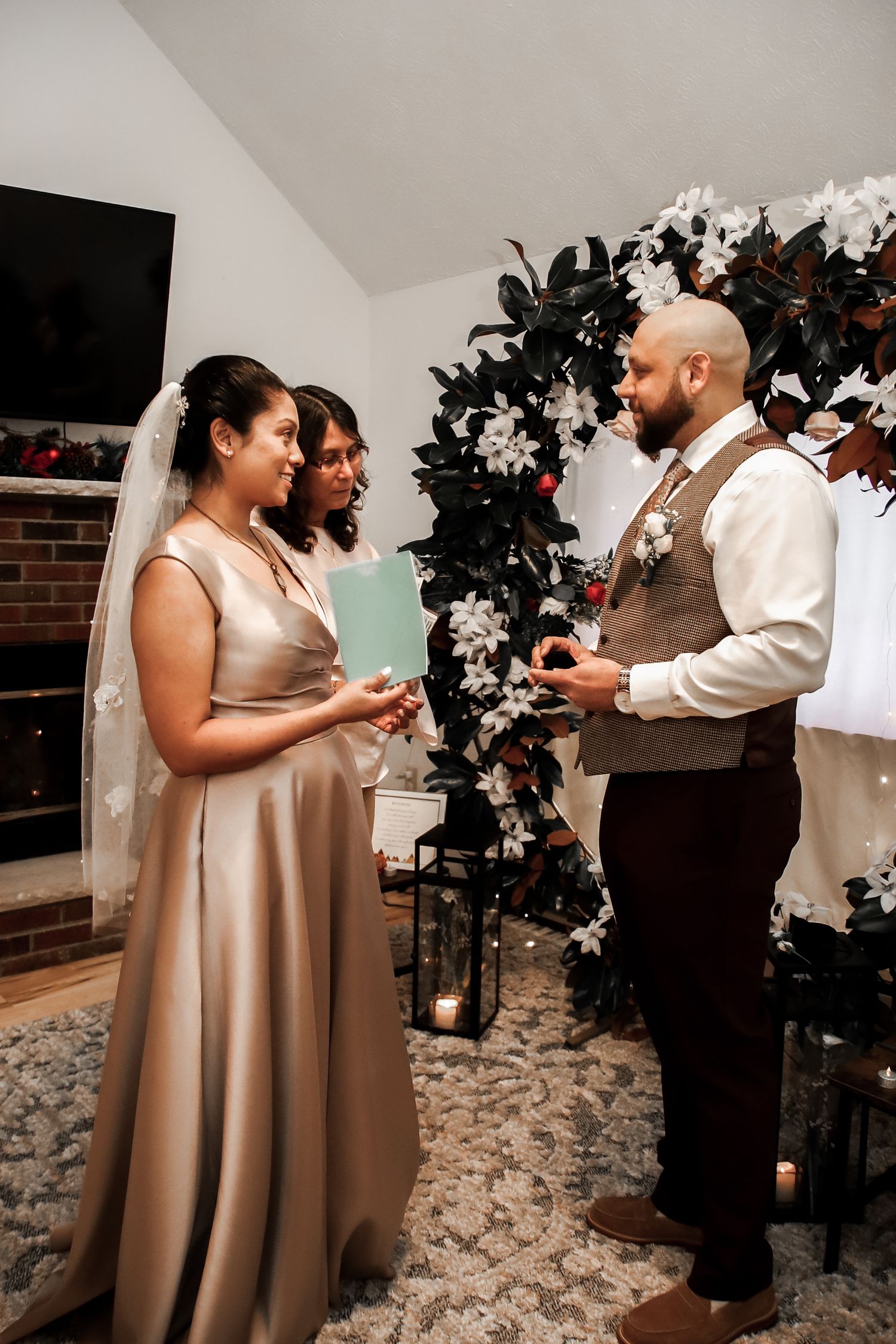 A wedding ceremony takes place under a floral arch with dark foliage and white flowers on a textured carpet.