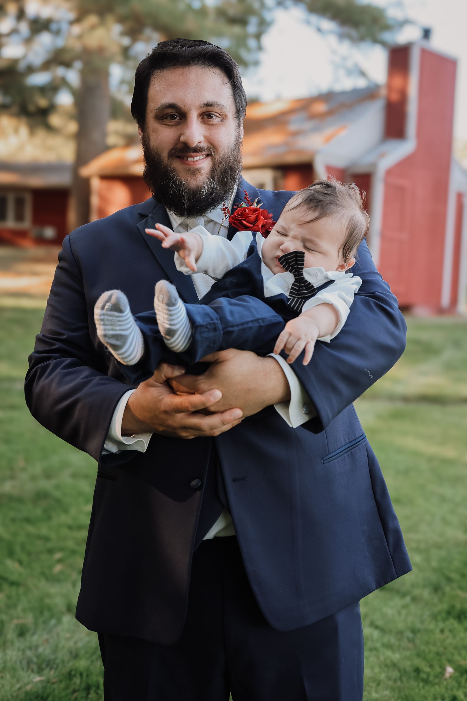 A man in a navy suit holds a laughing baby dressed in formal wear with a red boutonniere at an outdoor event.
