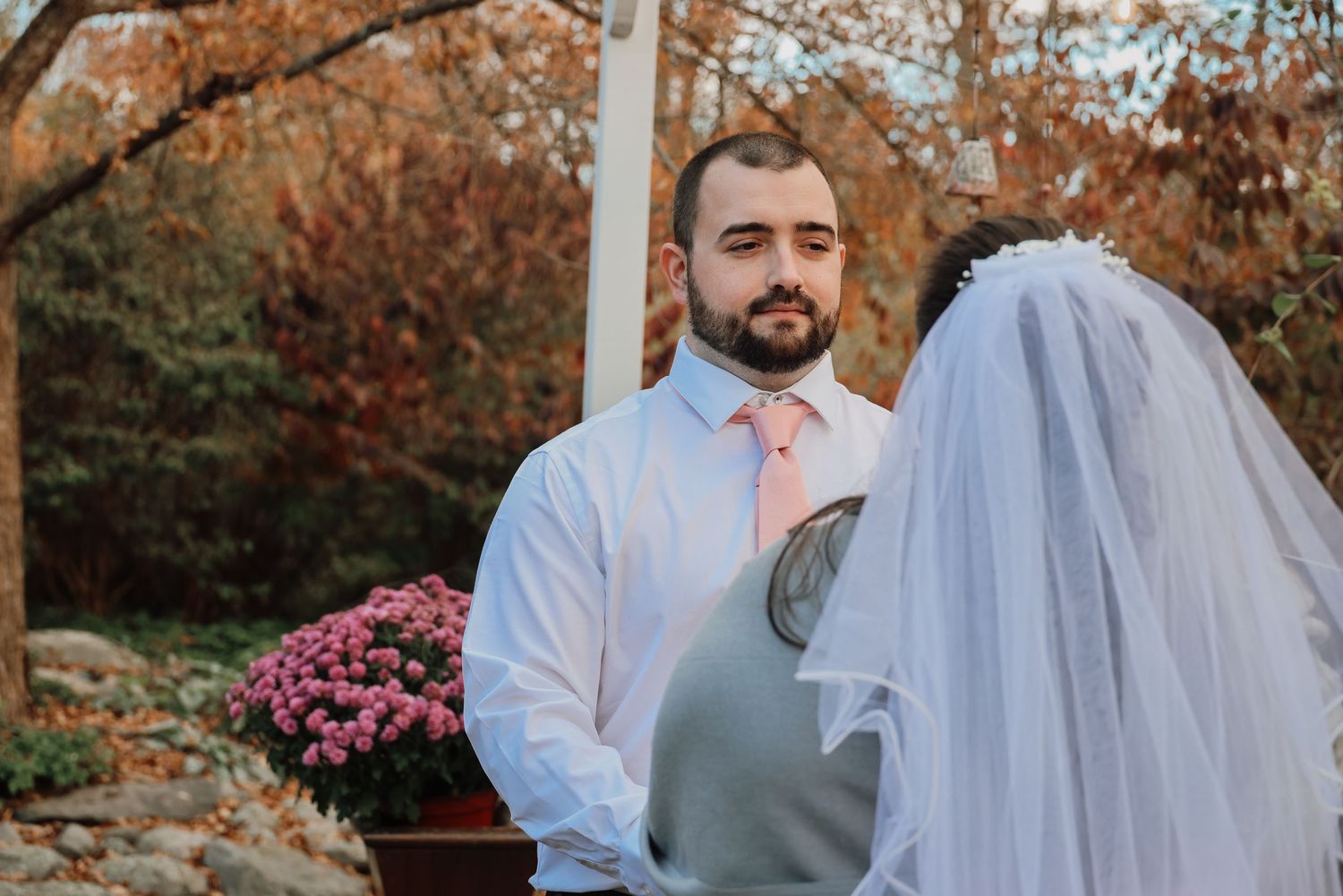 A couple in wedding attire embraces in a garden setting with autumn foliage and pink flowers in the background.
