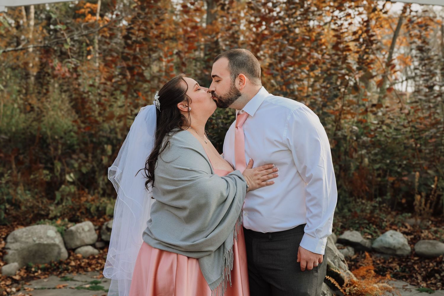 A couple shares a romantic moment in front of autumn foliage wearing soft pastel colors.
