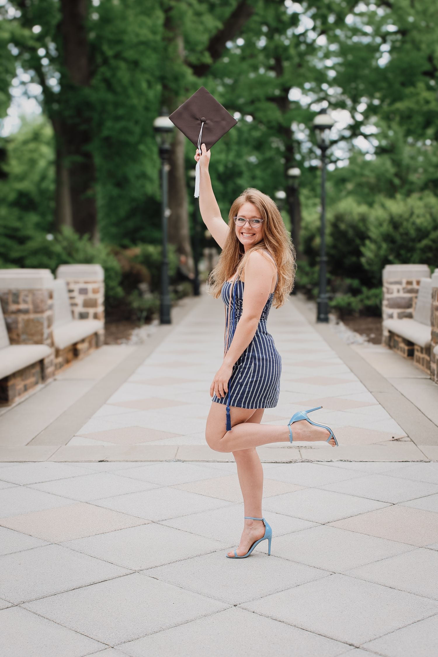 A graduate in a striped dress and blue heels poses on a stone walkway while holding up their graduation cap.