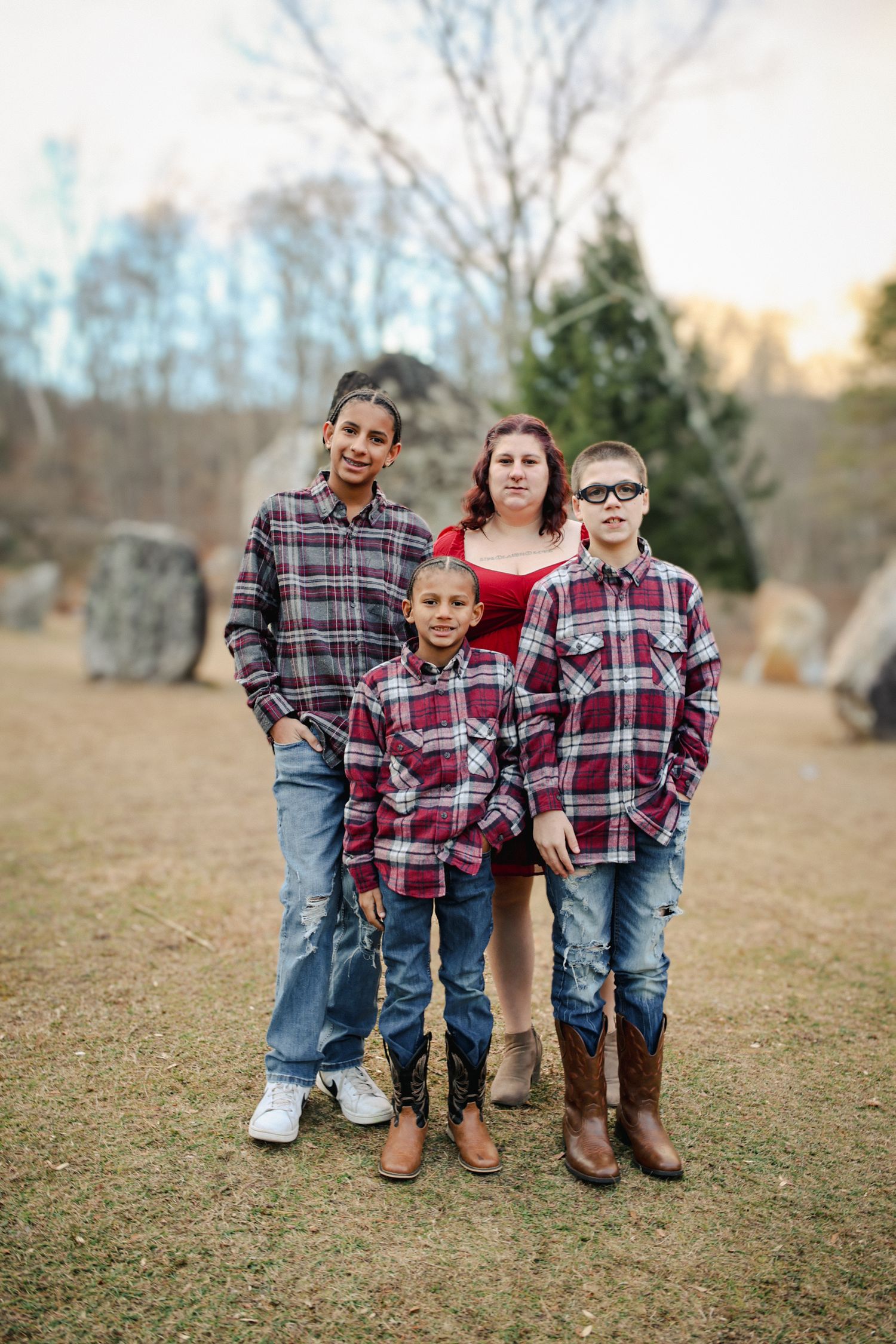 Three children wearing coordinating plaid shirts and jeans pose together on grass during an outdoor photo session.