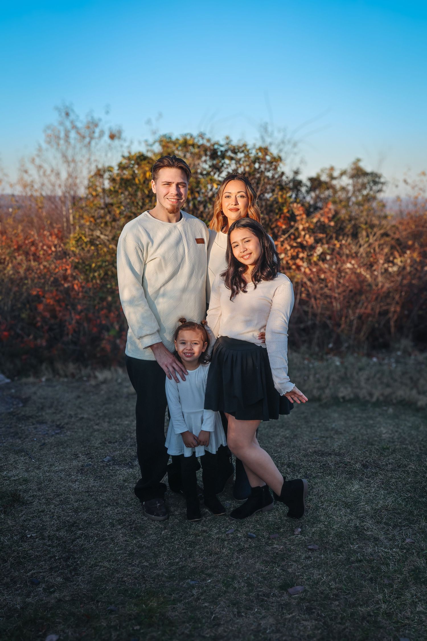 A family poses together outdoors at sunset with autumn foliage in the background.