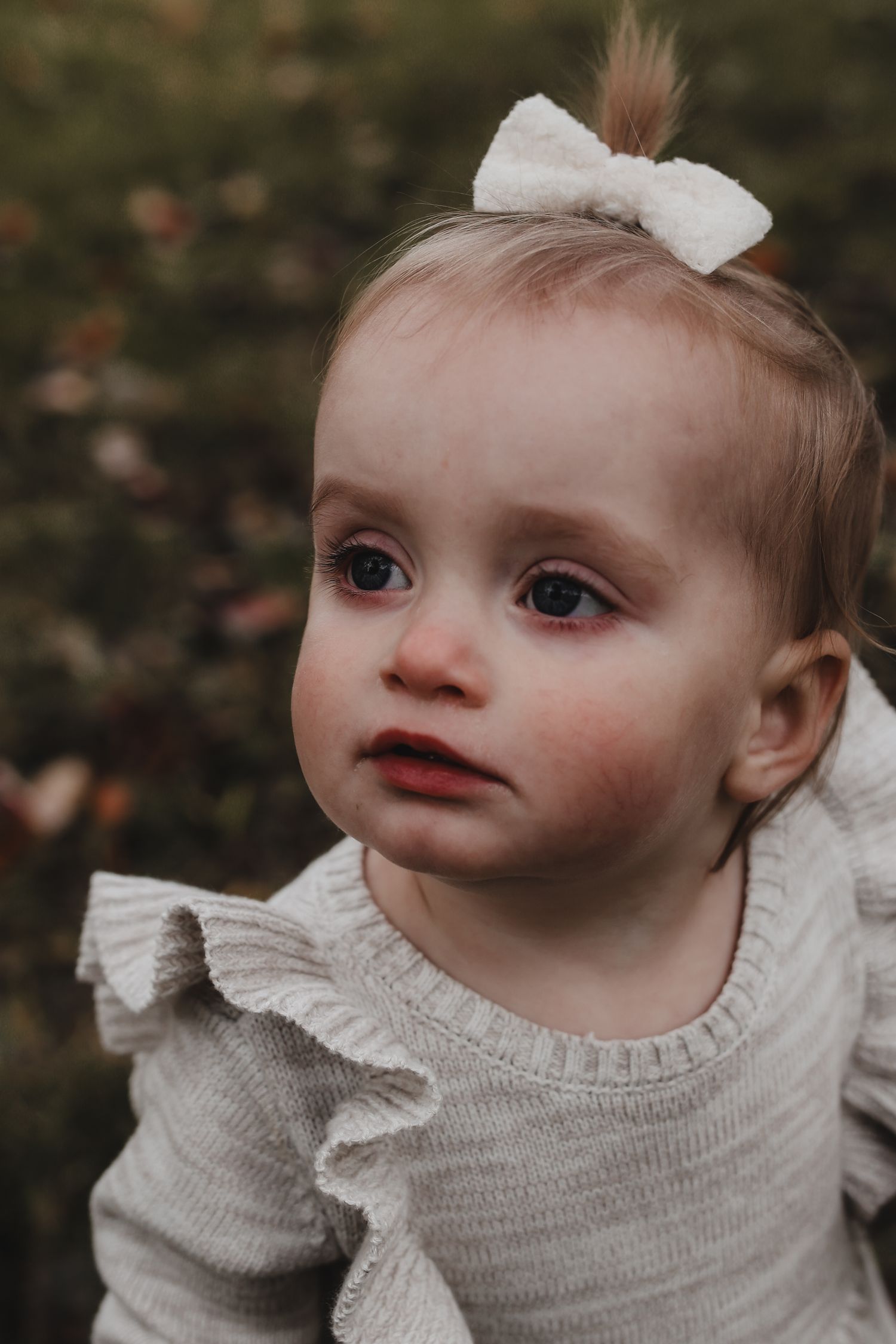 A young child wearing a white bow and beige ruffled sweater looks off to the side outdoors.
