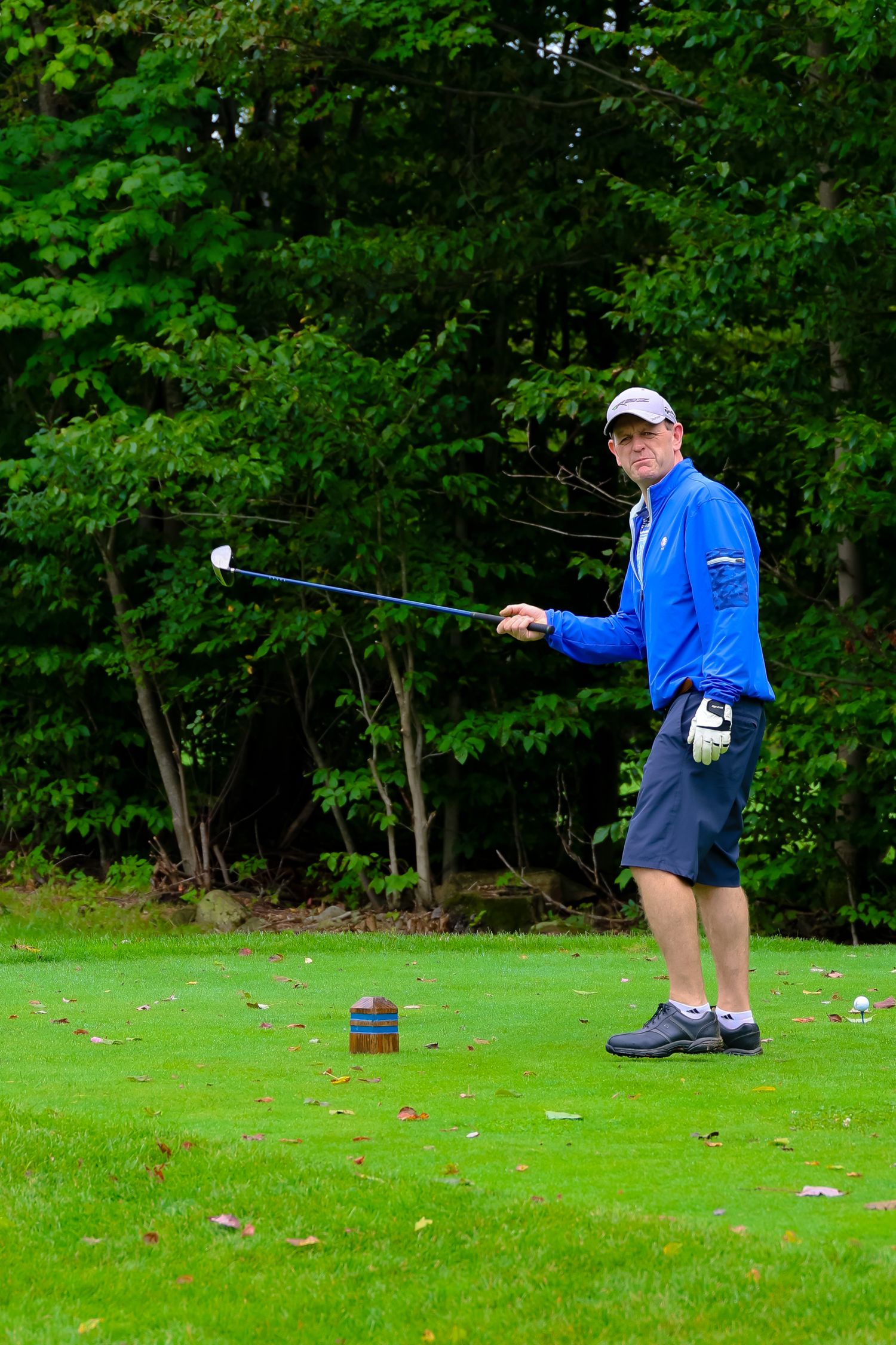 A golfer in a bright blue jacket takes a practice swing on a lush green tee box surrounded by trees.