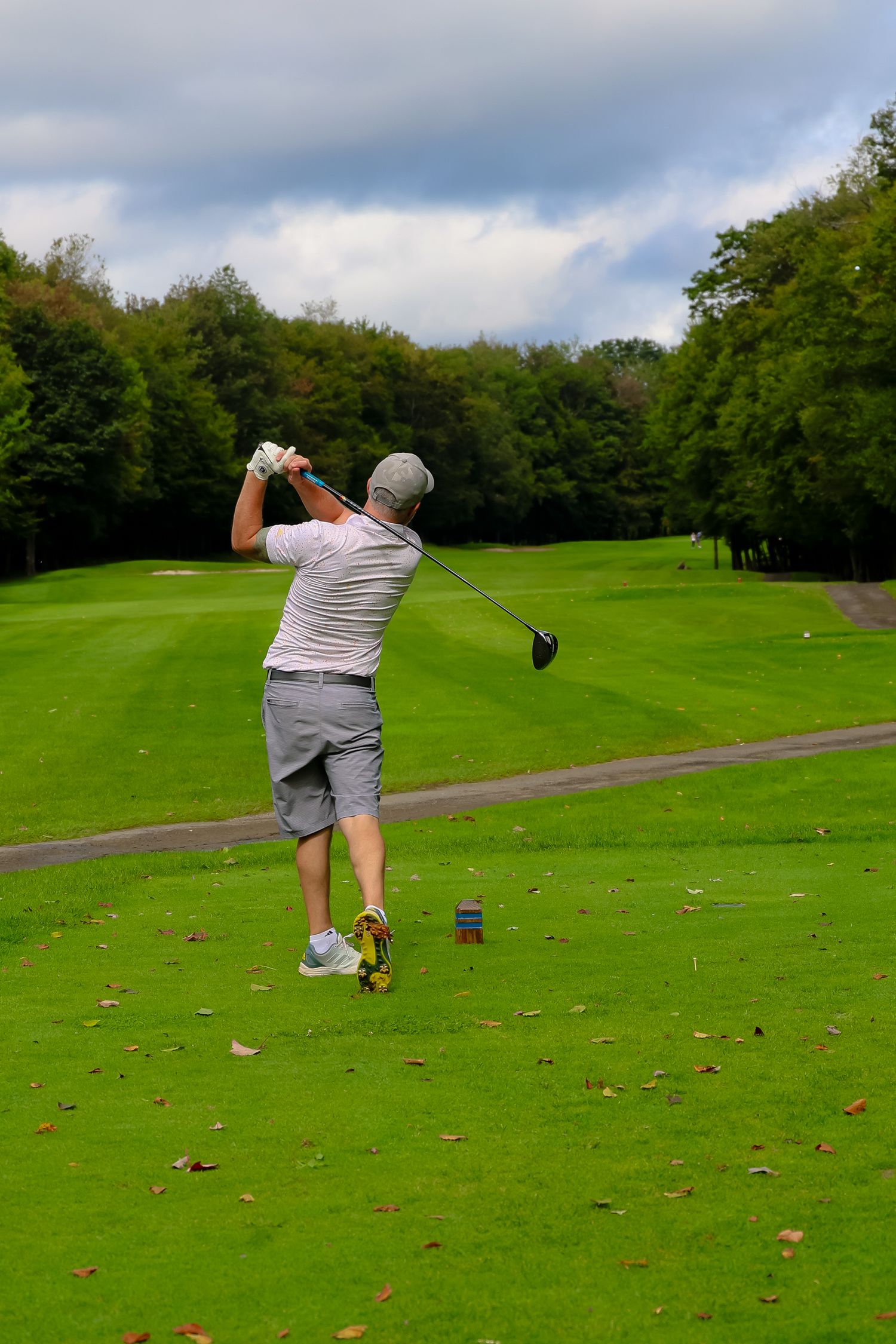 Golfer in grey attire takes a swing from the tee box on a lush green golf course surrounded by trees.