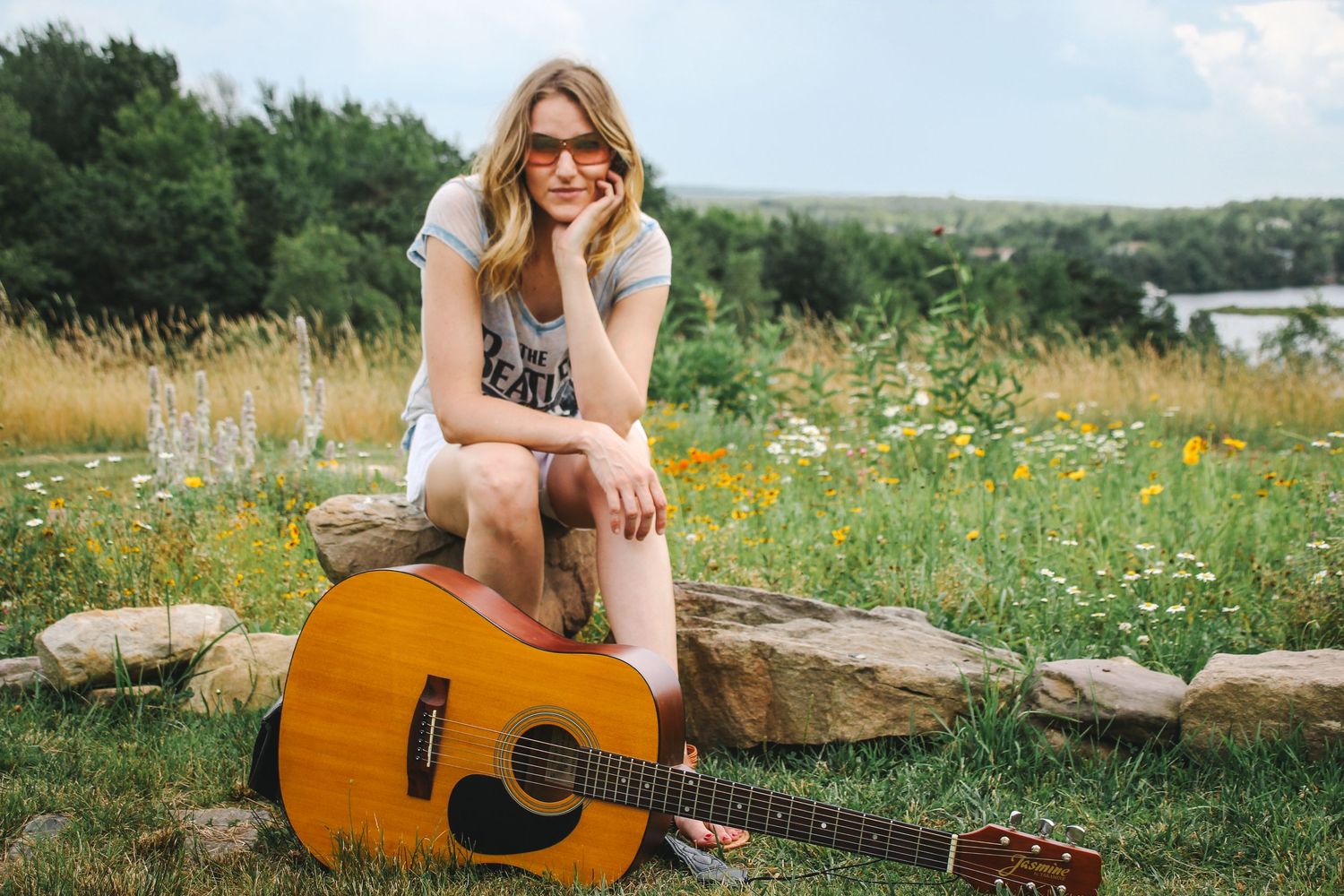 A guitar player sits in a wildflower meadow on a sunny day with an acoustic guitar and sunglasses.