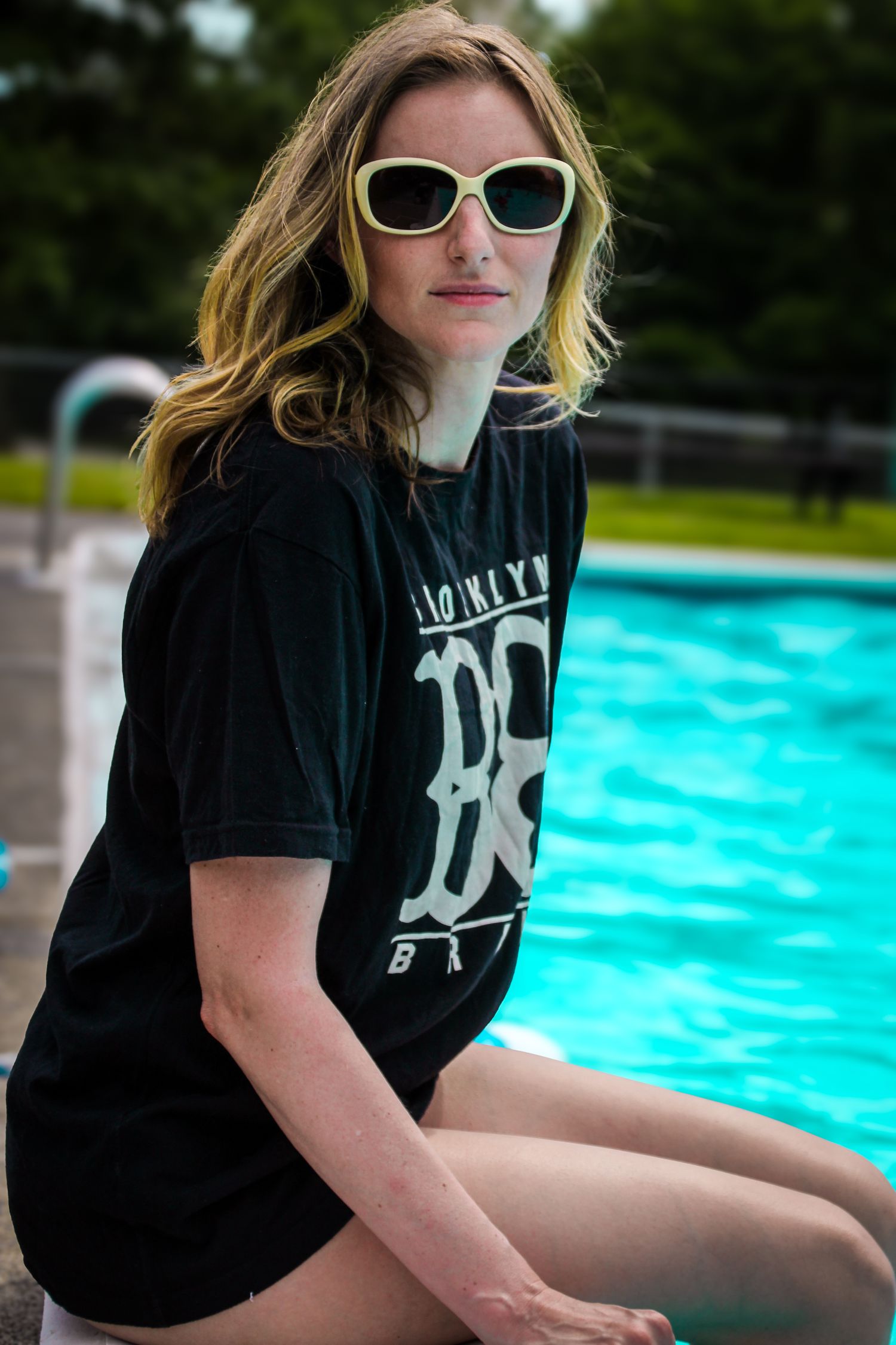 Person in sunglasses and black t-shirt poses near bright blue swimming pool in outdoor setting.