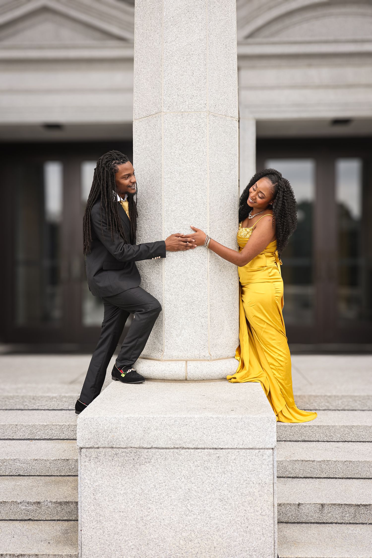 A couple in formal attire playfully poses on white stone steps, with one person in yellow and one in black.