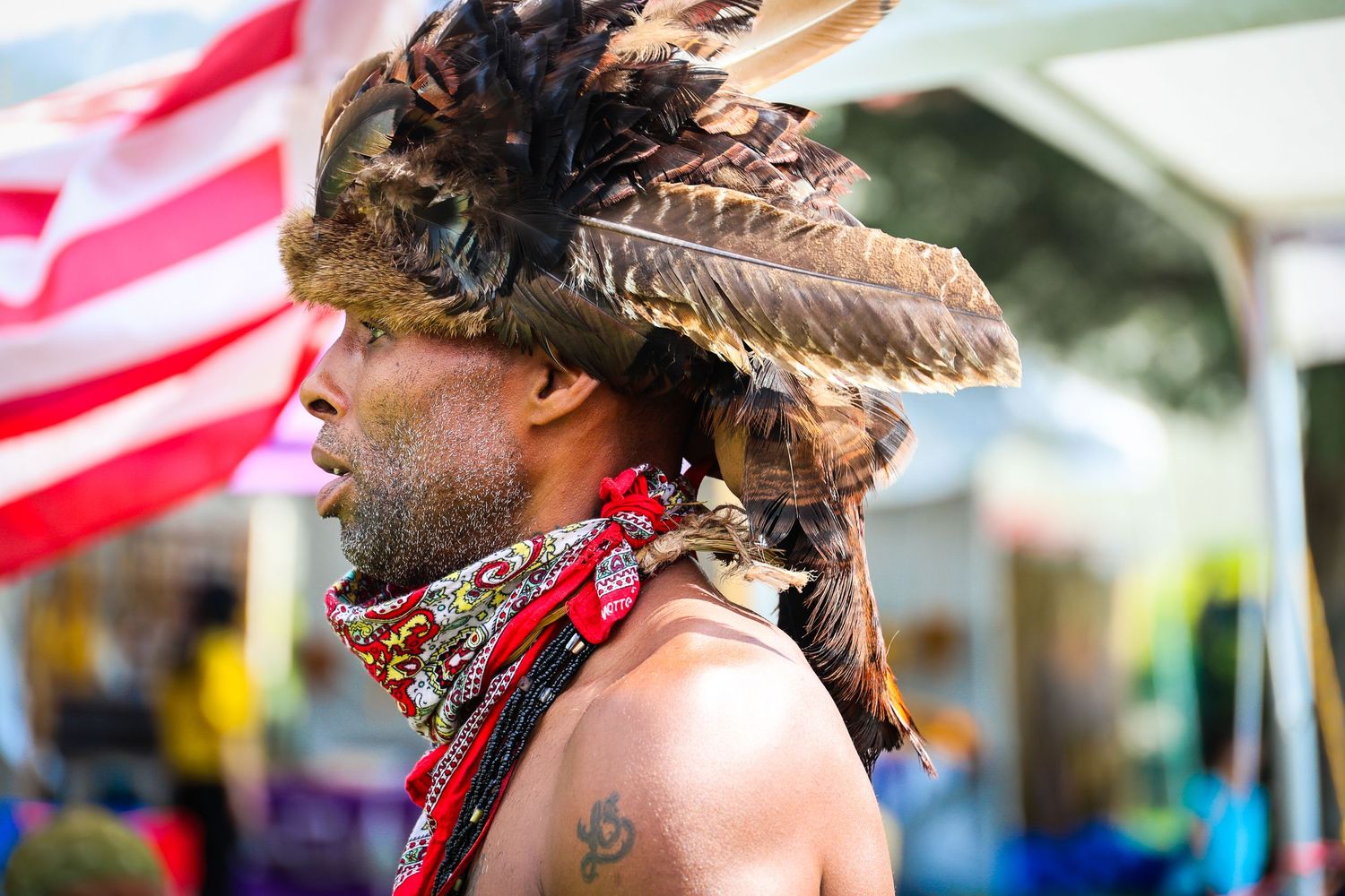 Native American dancer wearing traditional feathered headdress and bandana performs at cultural festival with US flag in background.