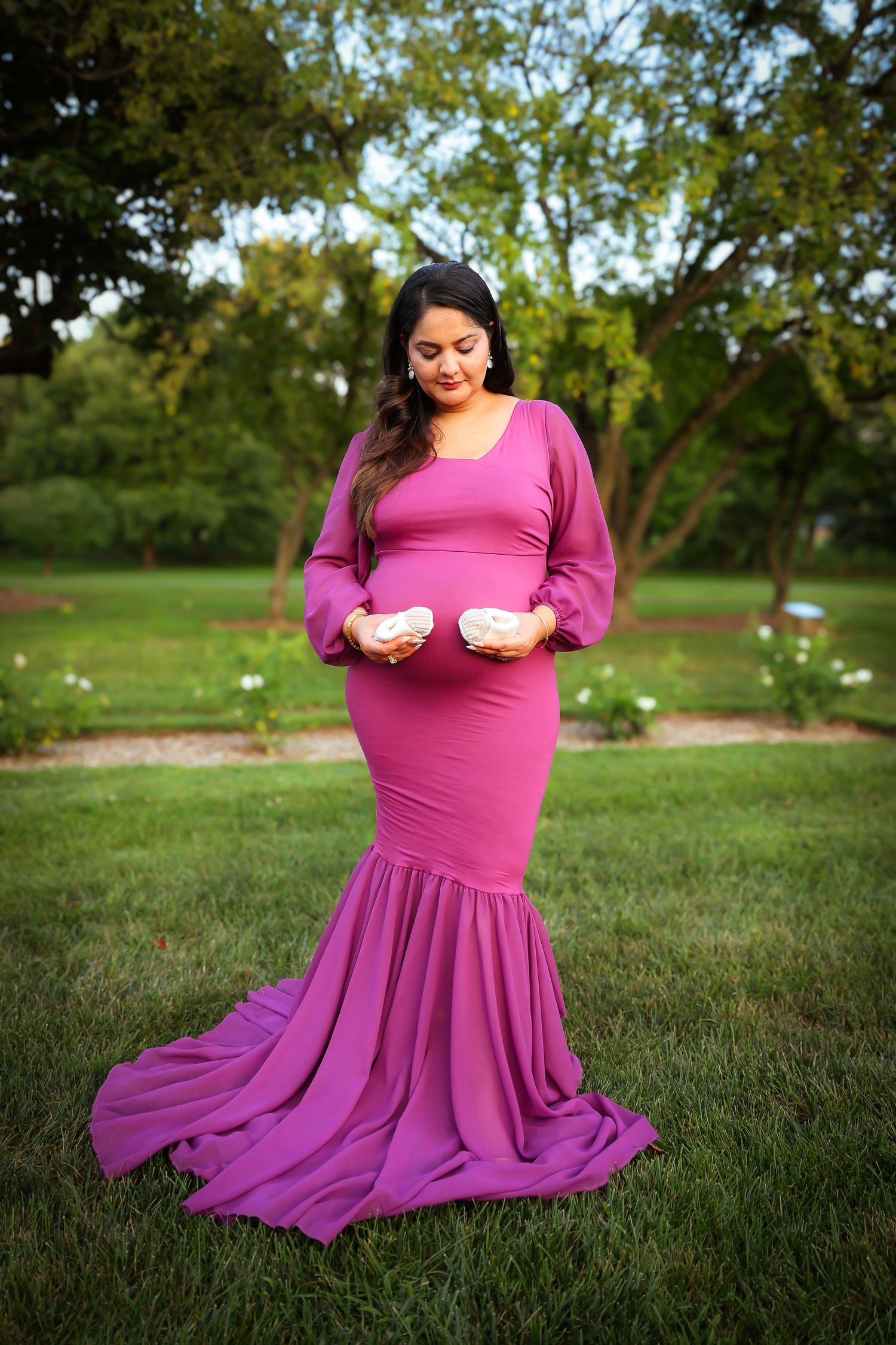 A person in a flowing magenta dress sits gracefully on green grass in a park setting during golden hour.
