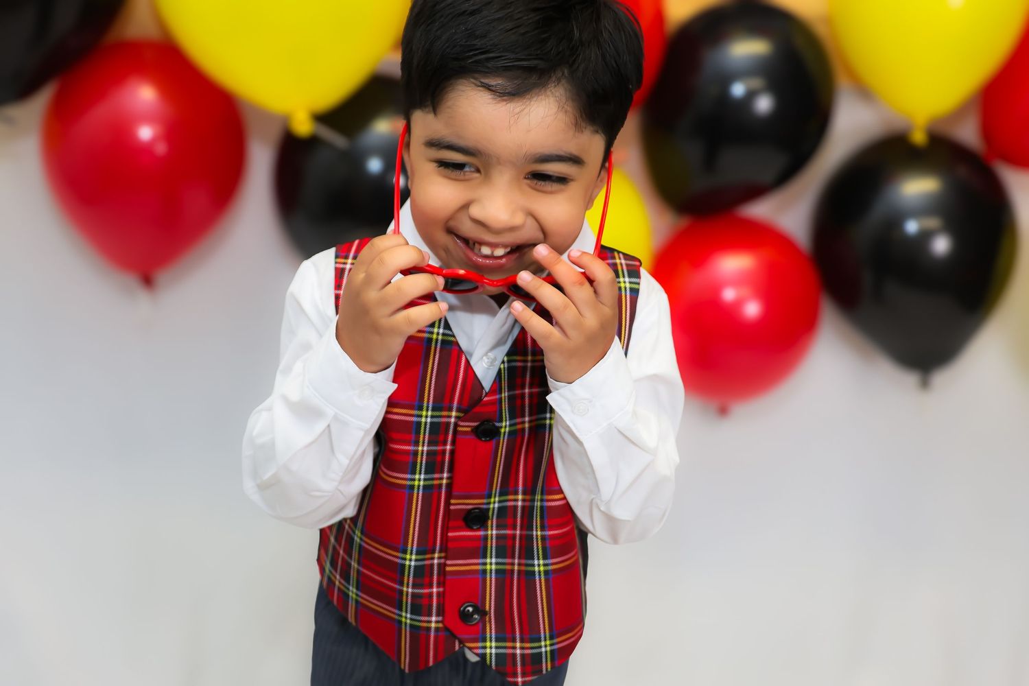 A young child in a red plaid vest and white shirt smiles while standing against a backdrop of yellow and red balloons.