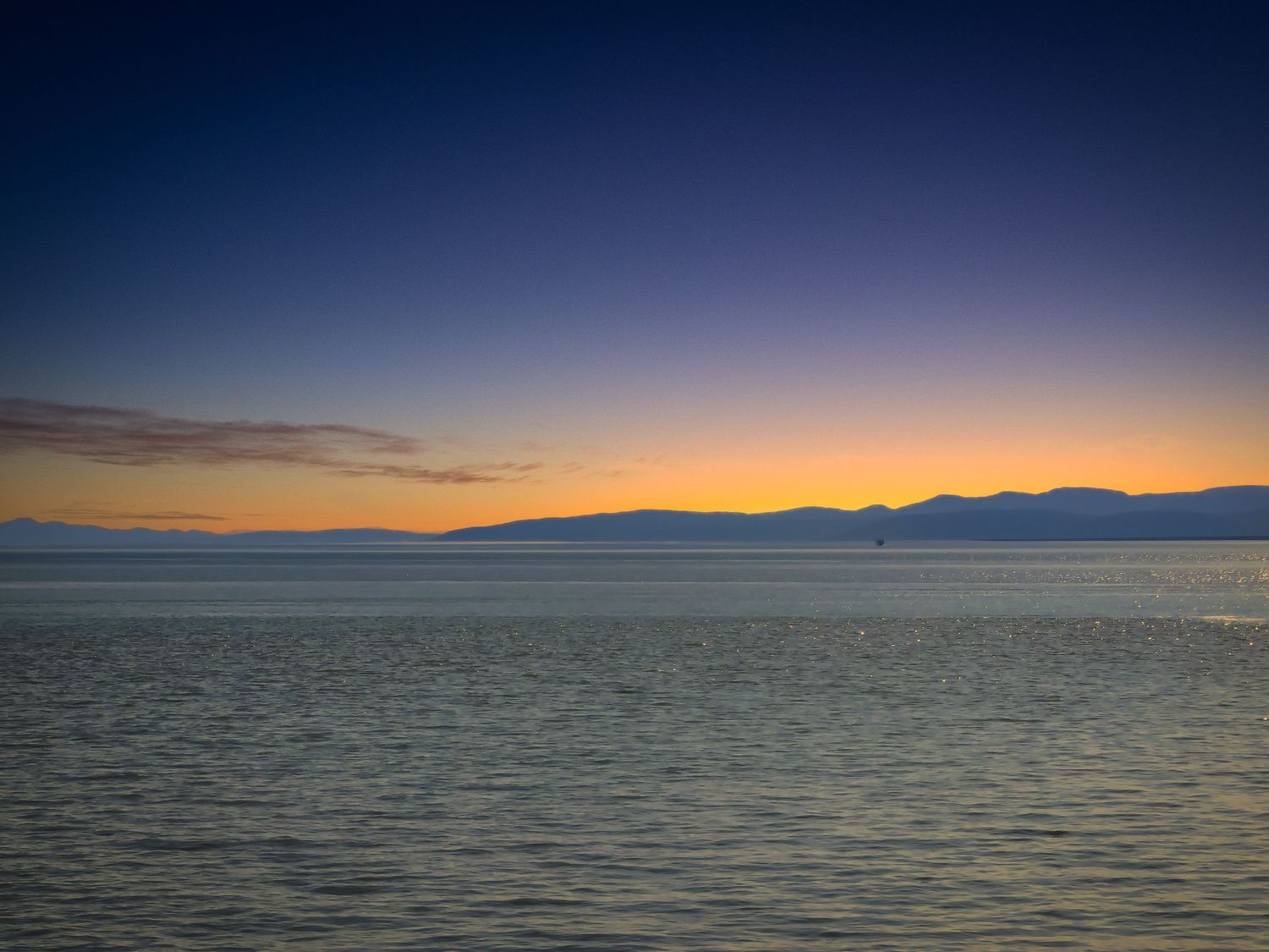 Tranquil sunset over a calm lake with mountains silhouetted against the darkening blue and orange sky.