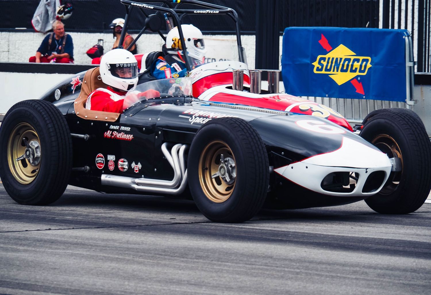 A vintage racing car with red and black livery speeds past a Sunoco sign at a motorsport event.