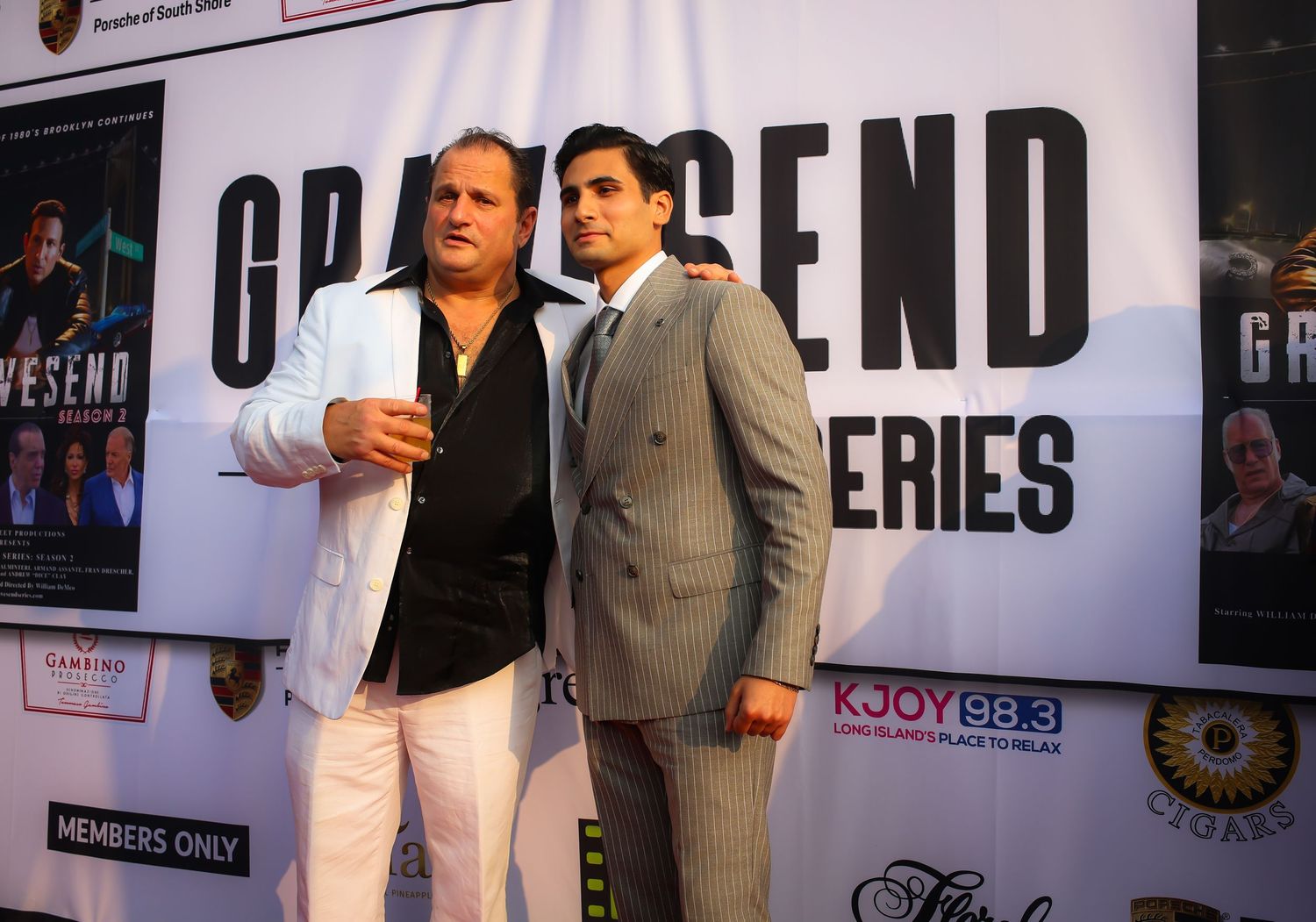 Two people in formal attire pose together at an event with promotional banners and signage in the background.