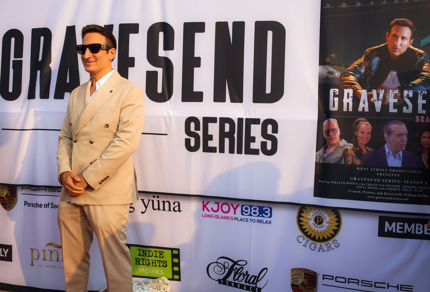 A person in a light colored suit and sunglasses poses on a red carpet in front of a Gravesend series backdrop.