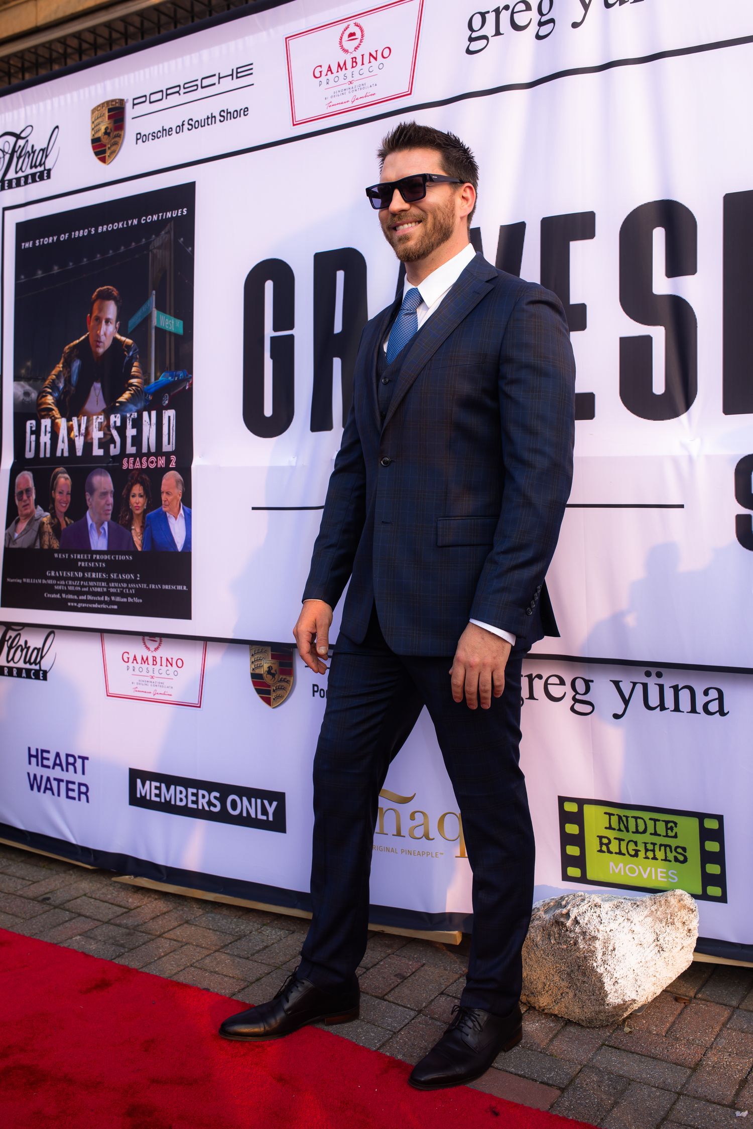 A figure in a dark suit stands on a red carpet in front of a step-and-repeat backdrop with sponsor logos.