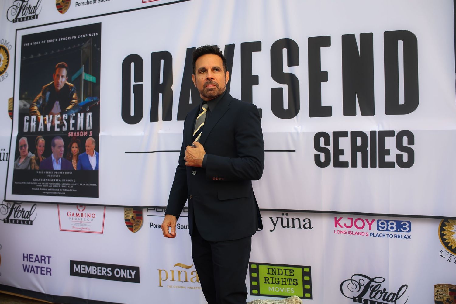 Person in black suit poses at red carpet event with Gracesend Series backdrop and sponsor logos.