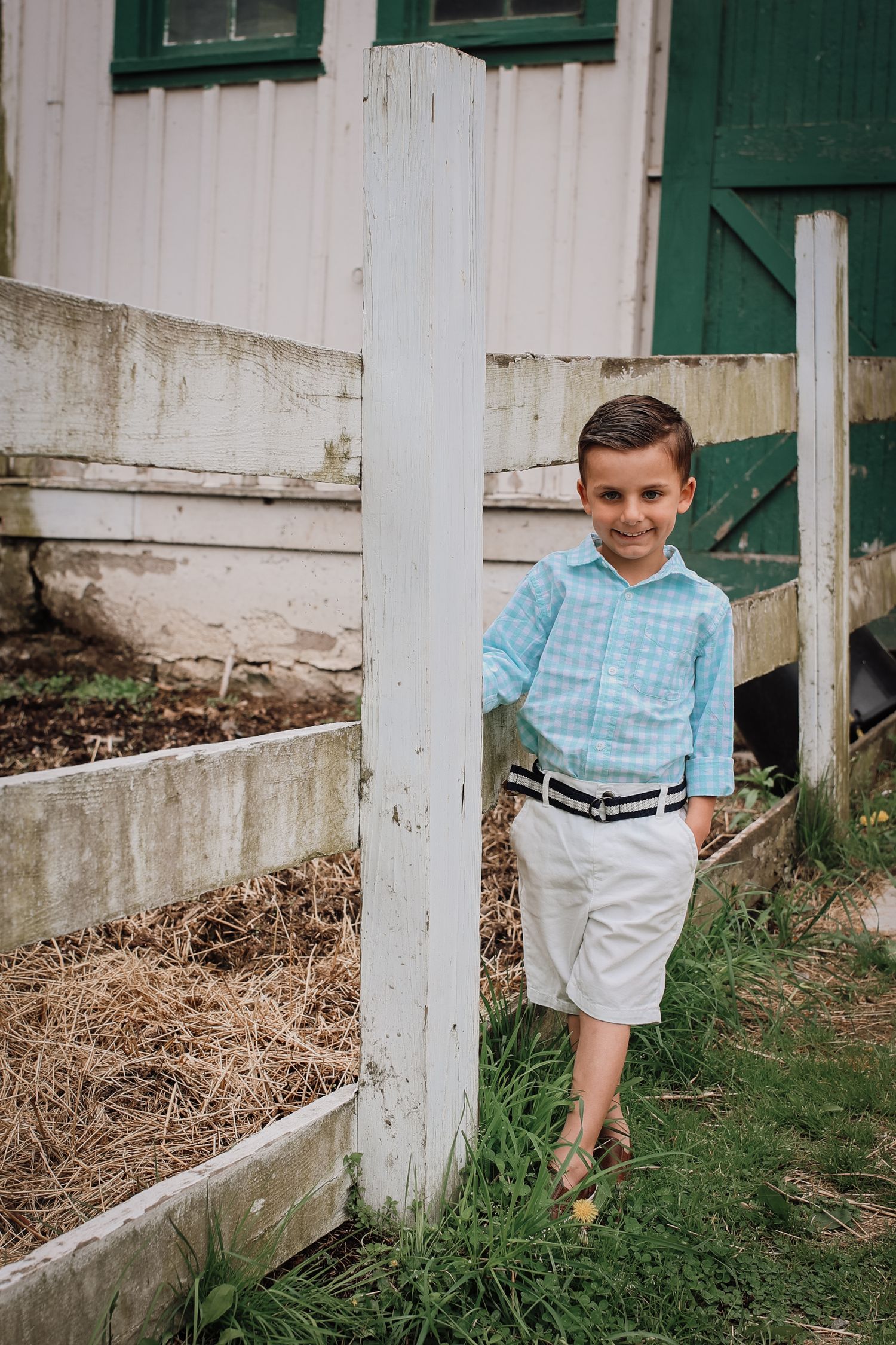 A child in a blue checkered shirt and white shorts stands playfully behind white fence posts in a rustic outdoor setting.