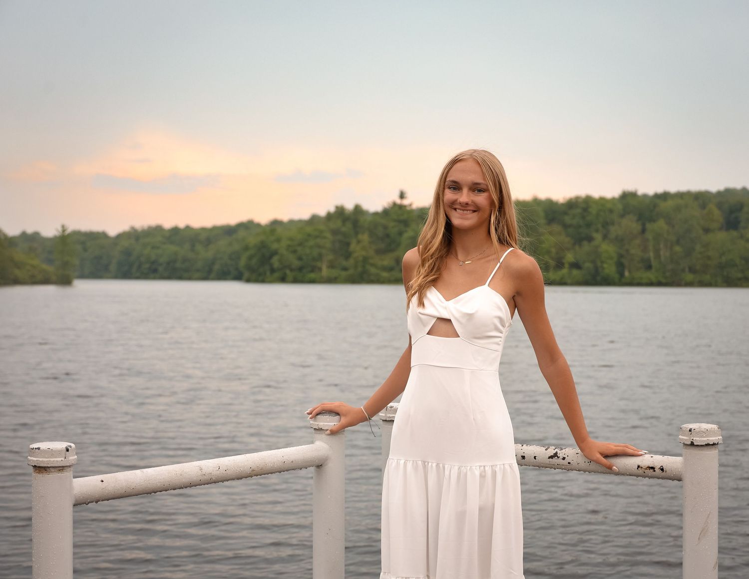 A person in a white dress stands on a wooden dock by a serene lake at sunset.