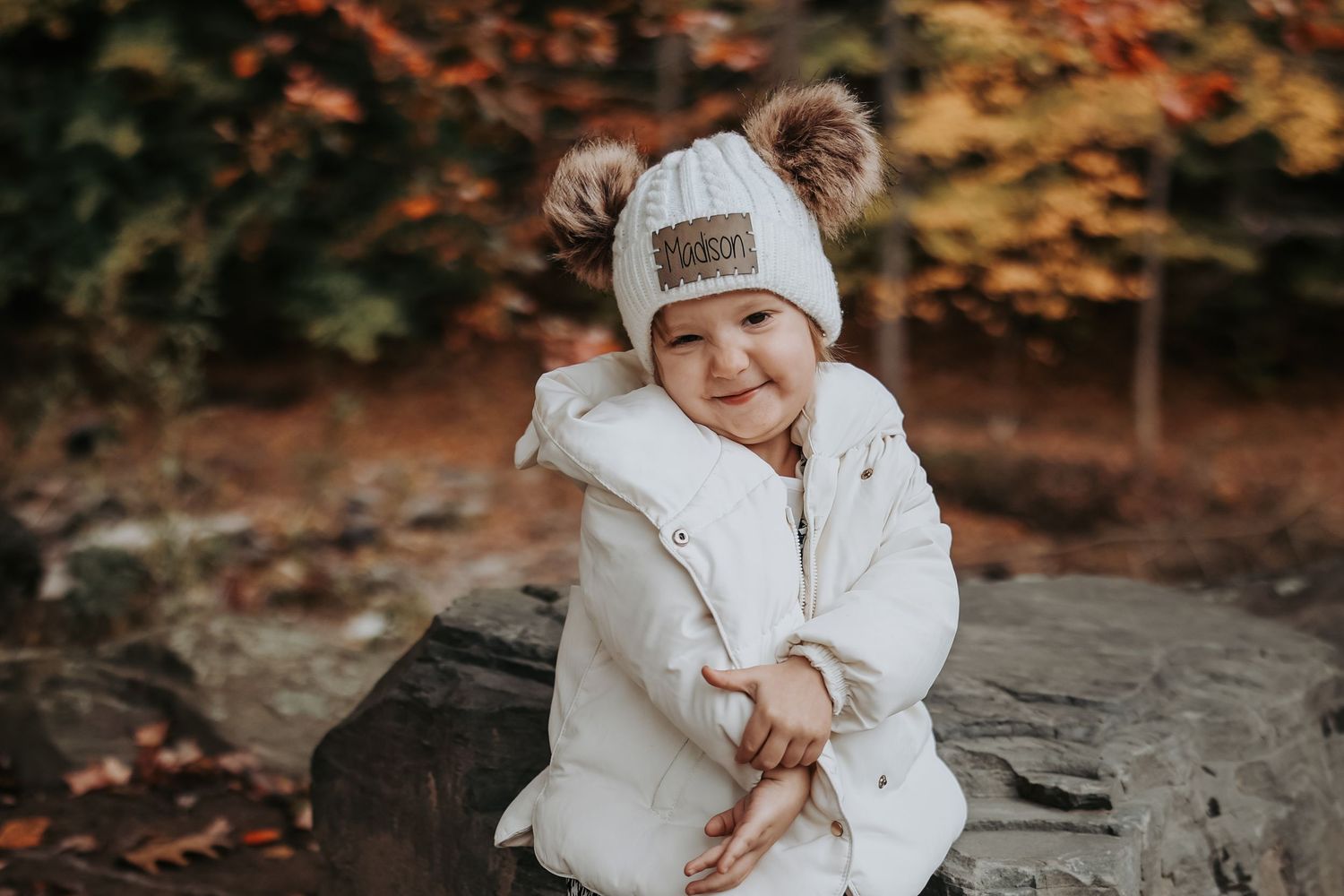 A baby wearing a white winter coat and beanie sits on a rock surrounded by autumn foliage.