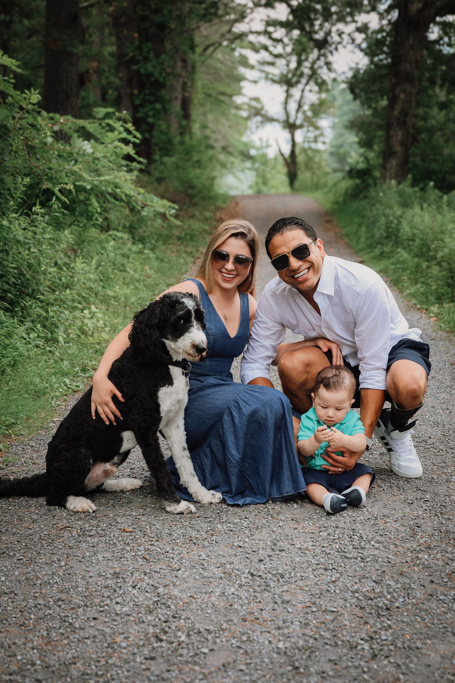 A family with their black dog sitting together on a gravel path in a lush green forest setting.
