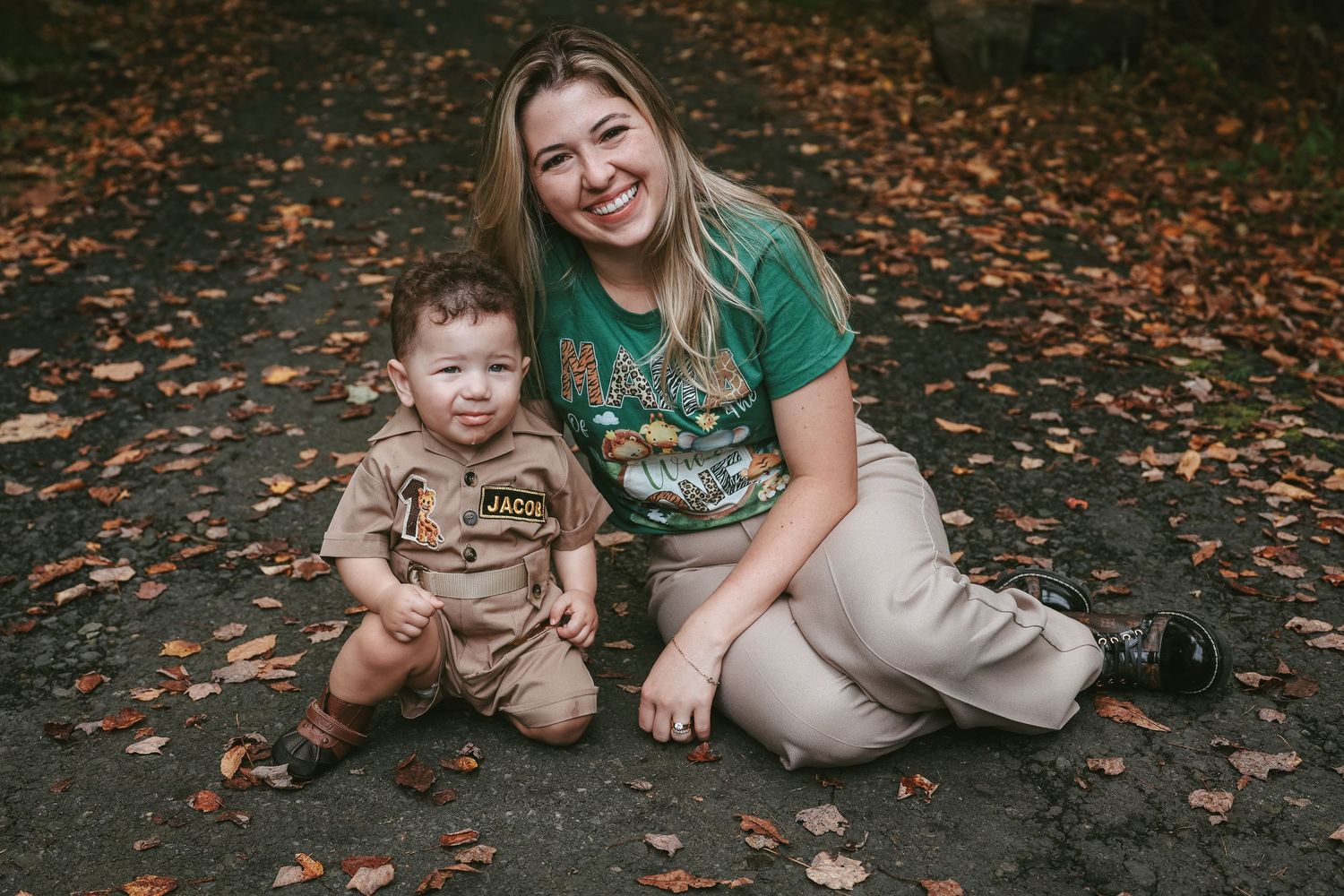 A parent and child sit together on a path covered in fallen autumn leaves in matching earth-toned outfits.