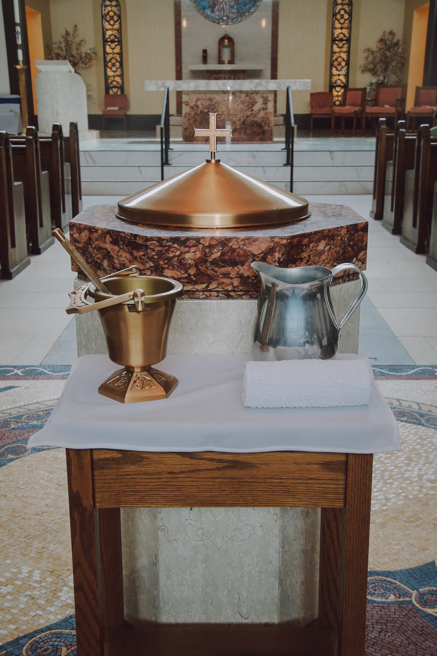 Brass church baptismal font and basin displayed on wooden pedestal with decorative mosaic floor in religious setting.