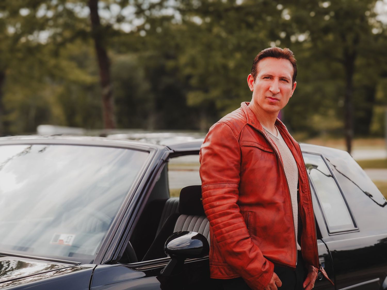 A person in a red leather jacket poses beside a black car in an outdoor setting.
