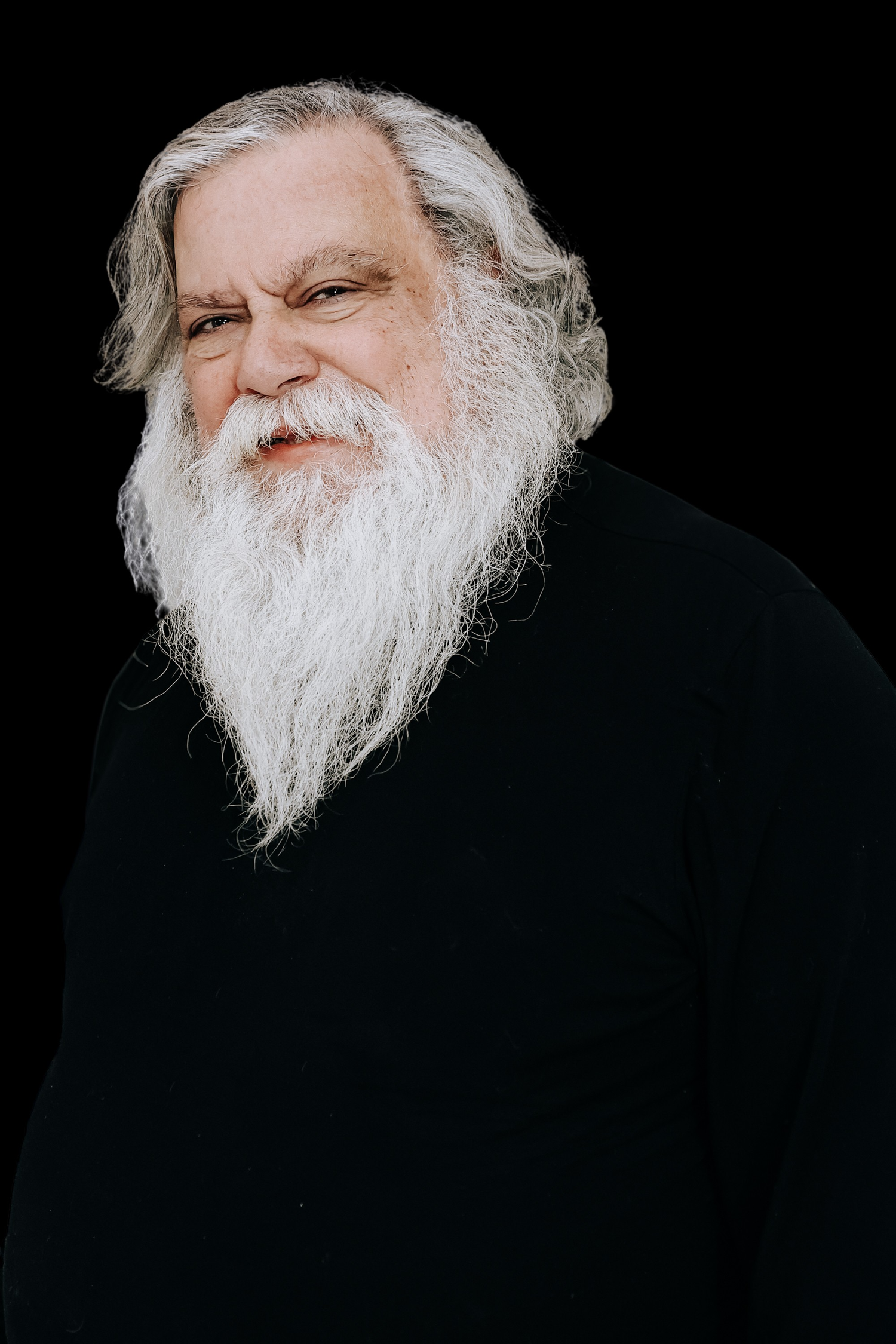 A person with a long flowing white beard is photographed against a dark background in dramatic lighting.
