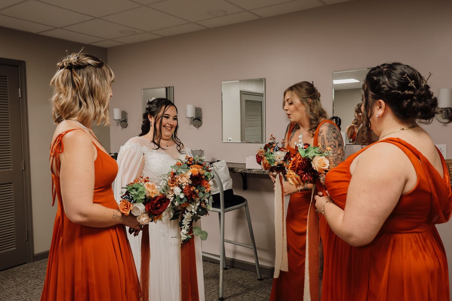 A group of bridesmaids in orange dresses share emotional moments and hugs before a wedding ceremony.
