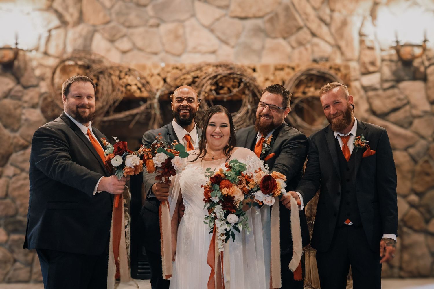 Wedding party laughs together in front of rustic stone wall with autumn floral bouquets.