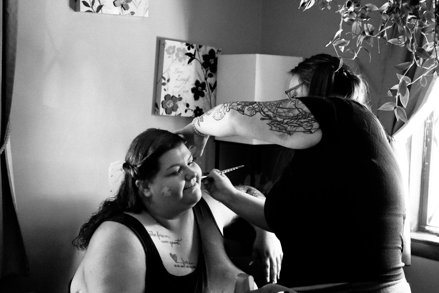 A makeup artist applies cosmetics to a client in a black and white photograph by a window.