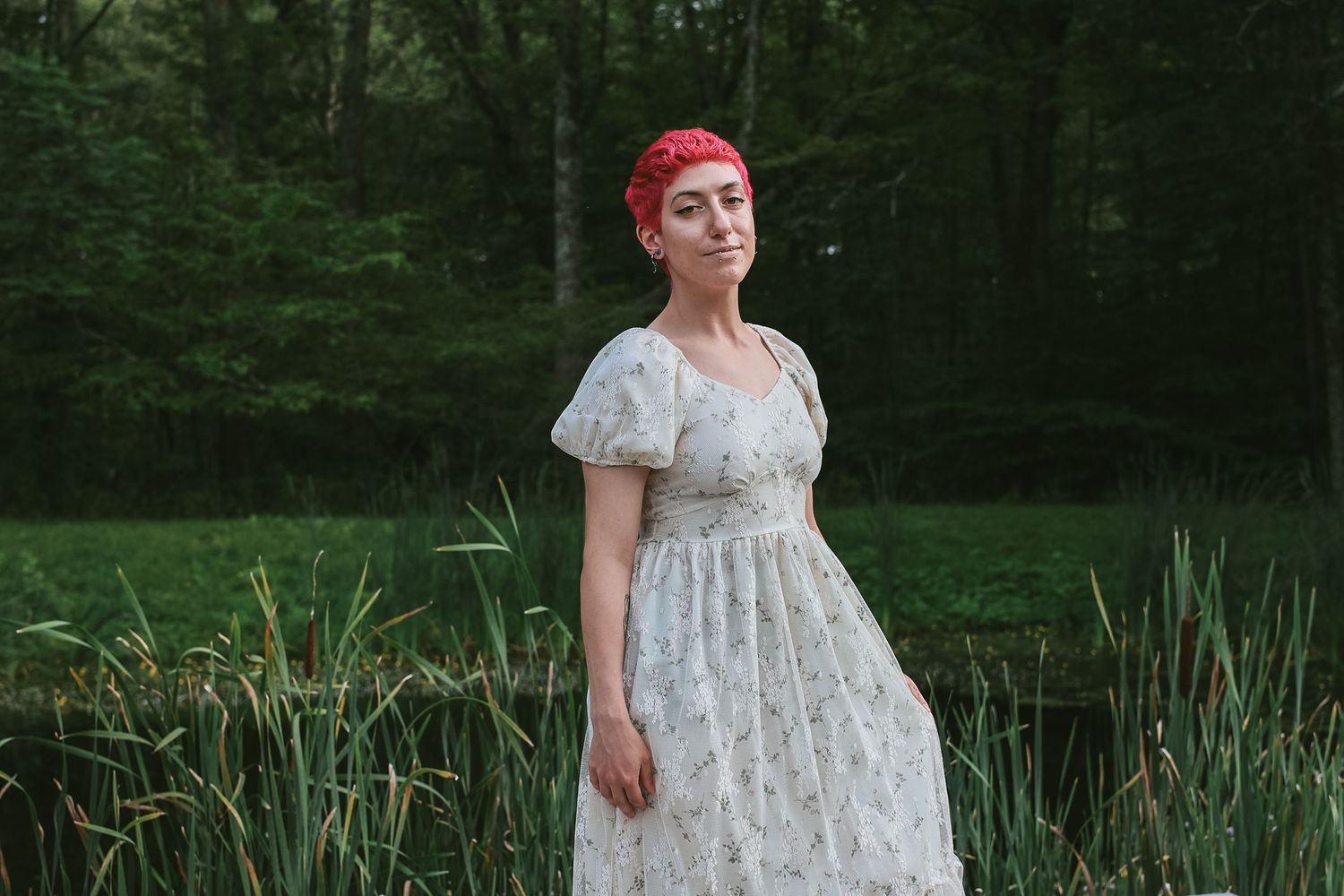 A person in a white floral dress poses in a lush green forest setting near a pond.