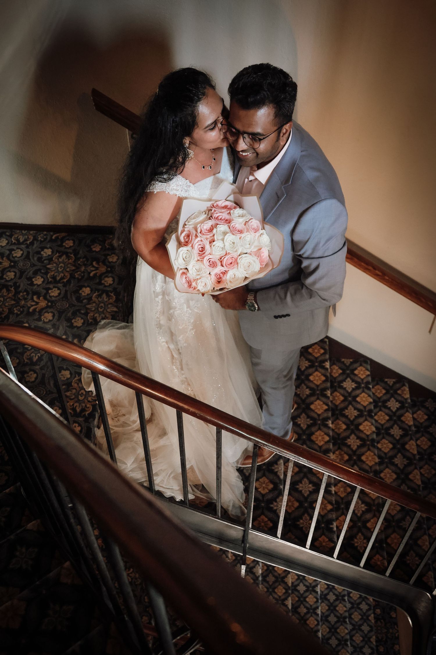 A couple in wedding attire shares a romantic moment on a staircase with patterned carpeting and wooden railings.