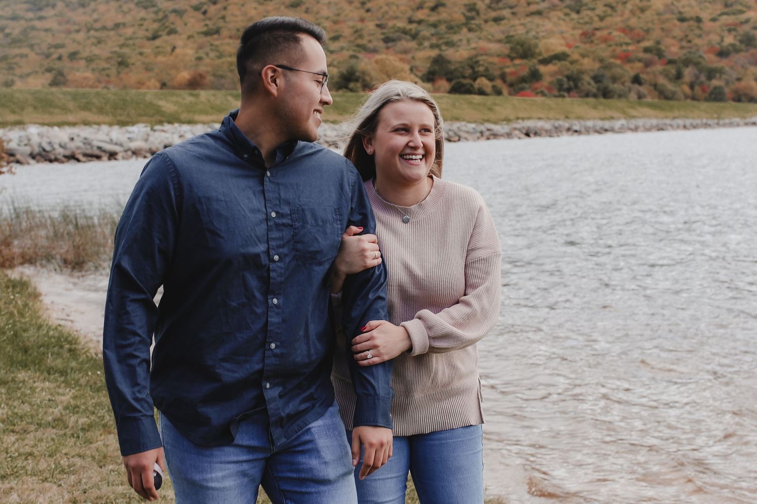 A couple walks and laughs together by a lakeside on an autumn day wearing casual sweaters and jeans.