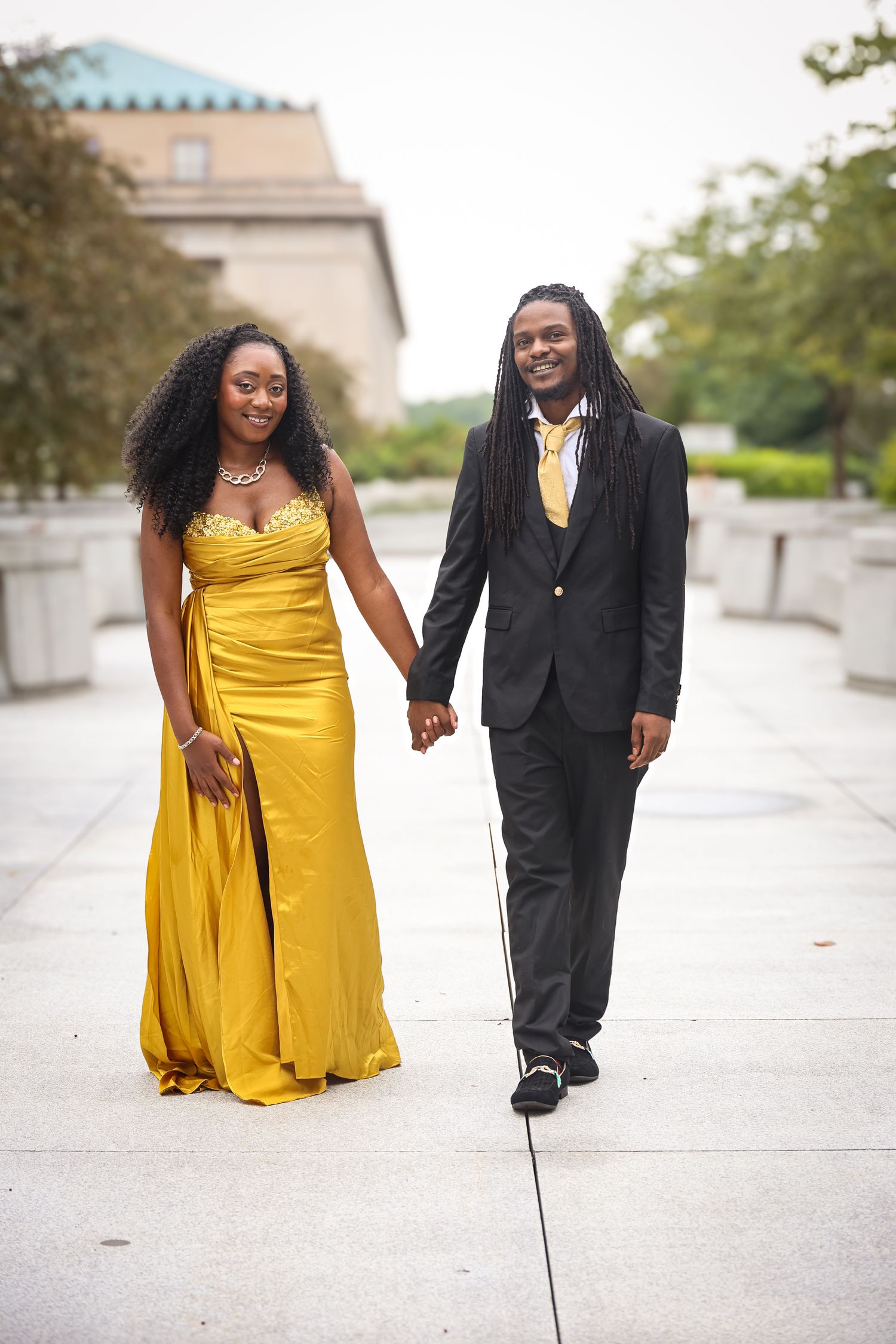 A couple in formal attire, with a yellow dress and black suit, walking together outdoors on a paved path.