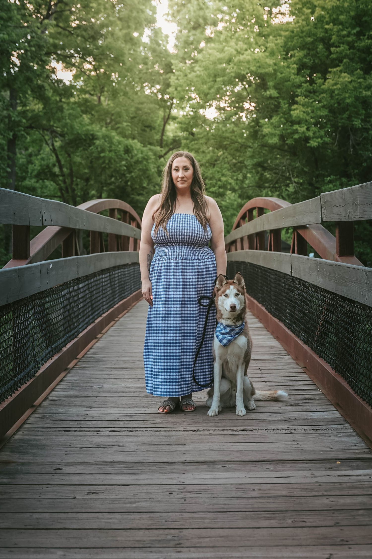 Woman in blue gingham maxi dress standing with Husky dog on wooden bridge surrounded by trees.