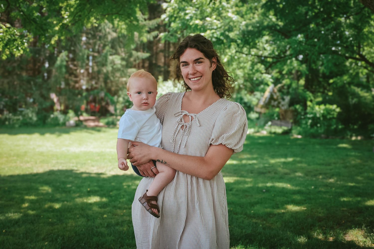 A smiling young woman in a beige dress holds a happy baby outdoors in a green garden setting.