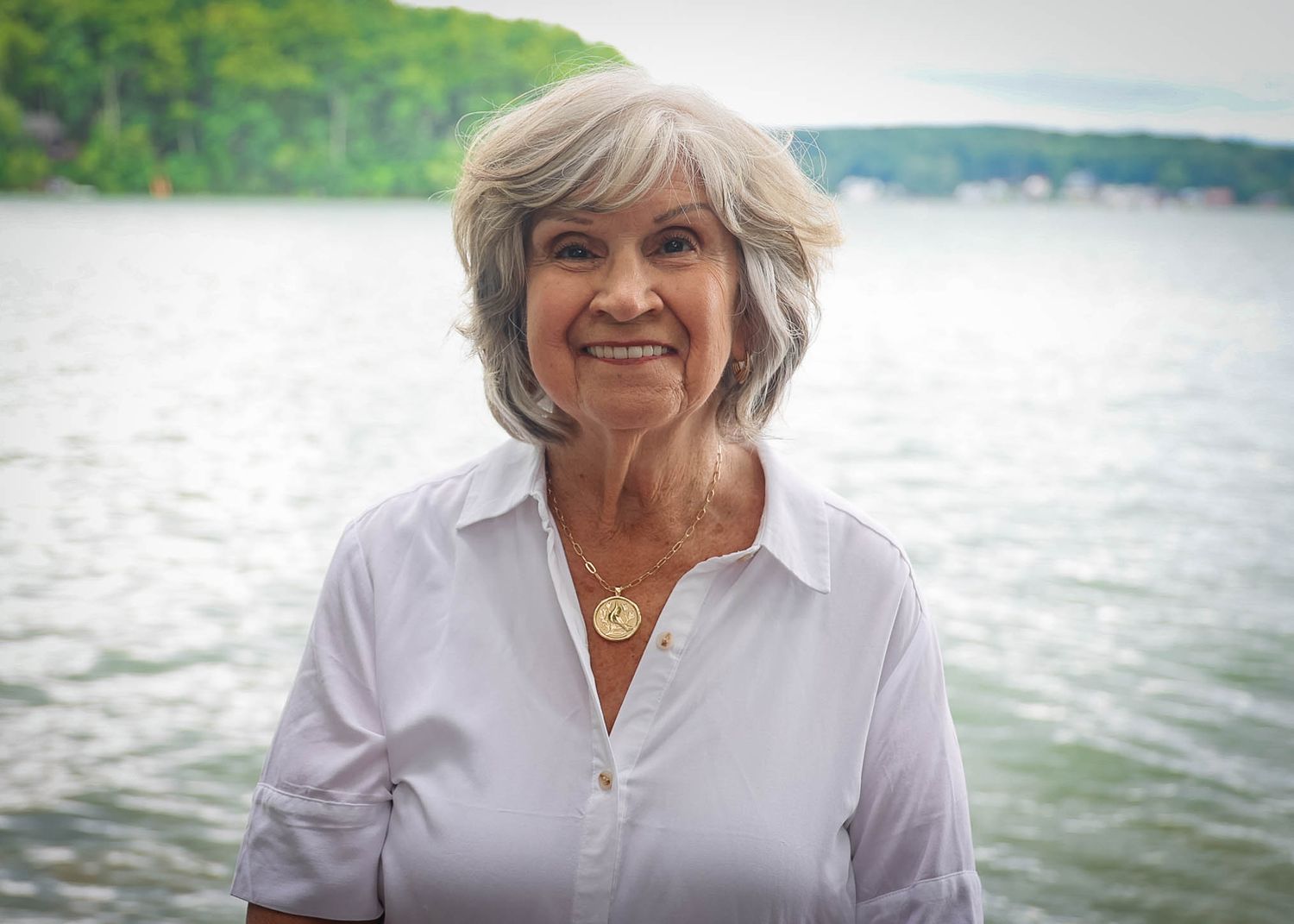 Smiling older woman with gray hair wearing a white shirt and gold necklace standing near a lake.
