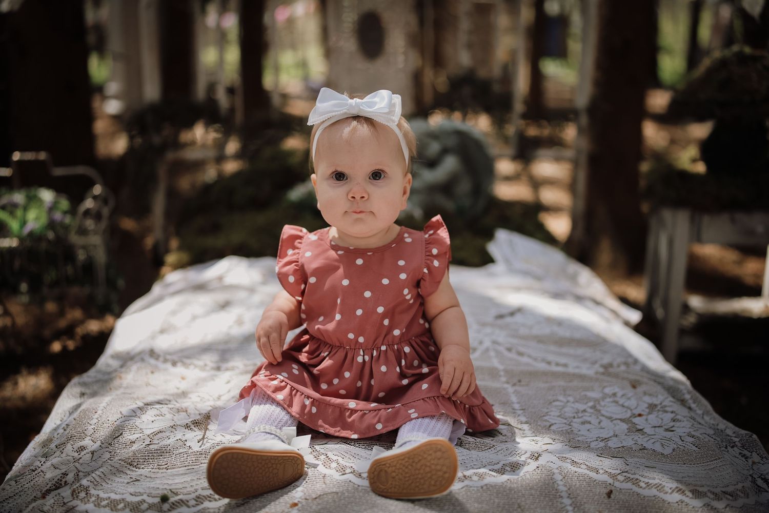 Baby girl in red polka dot dress with white bow headband sitting on lace blanket outdoors.