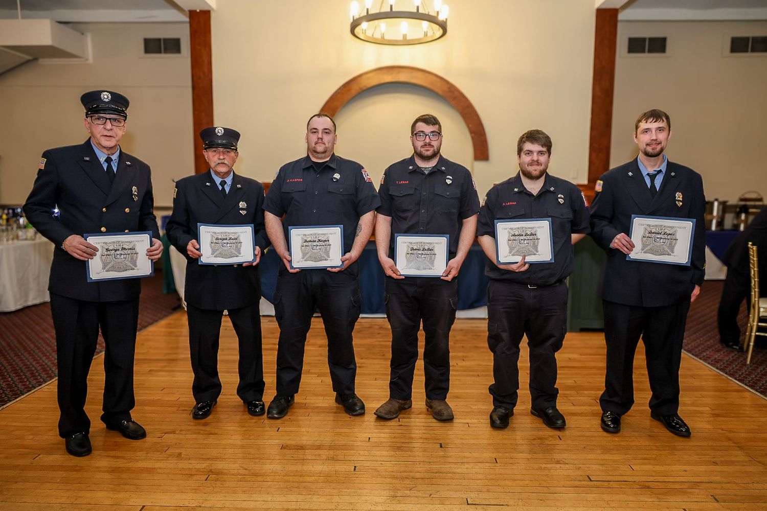 Six firefighters in dress uniforms proudly display certificates at an awards ceremony in a formal banquet hall.